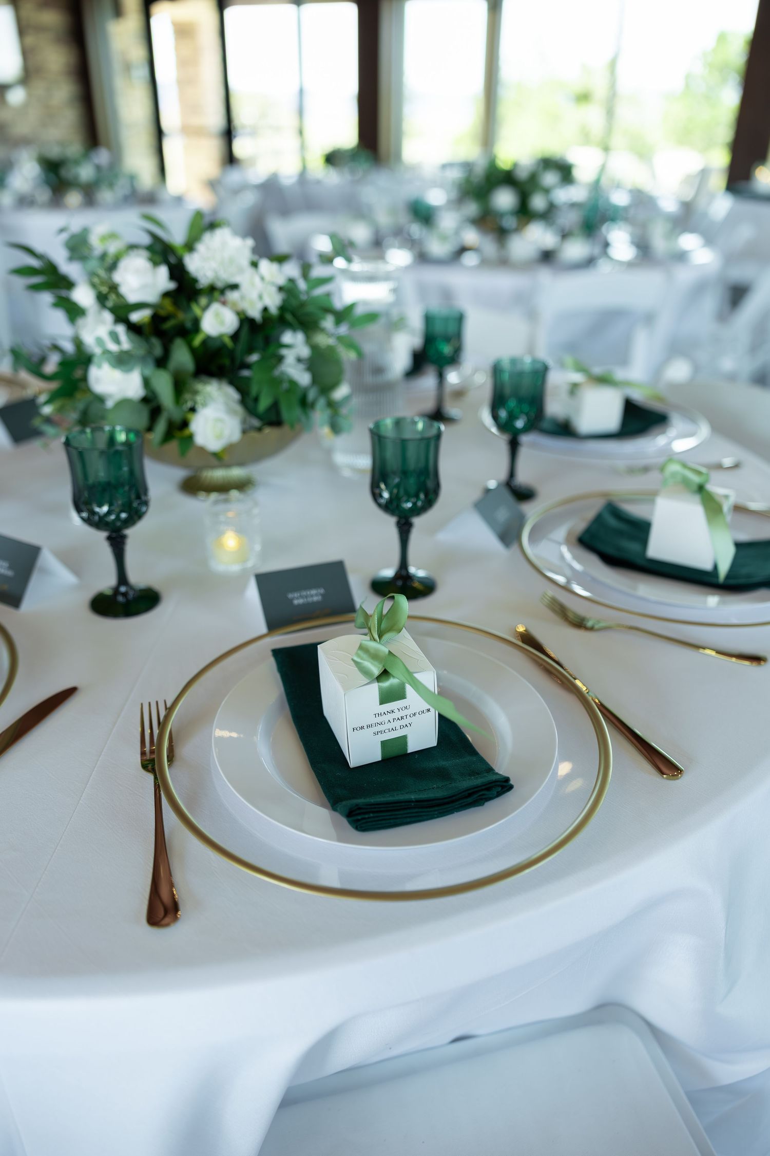 Elegant wedding reception table with green napkins, gold-rimmed chargers, emerald glassware, and white floral centerpieces.