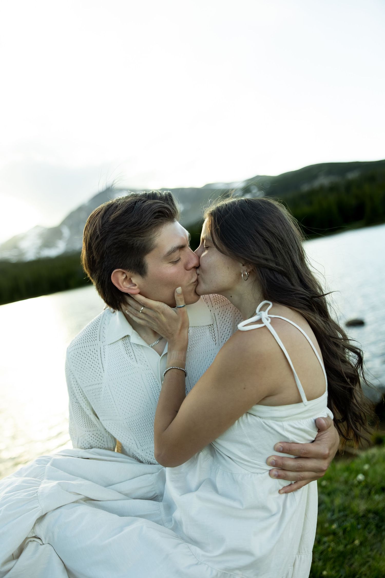 Couple sharing a romantic kiss by a mountain lake, both dressed in white, with snow-capped peaks in the background.