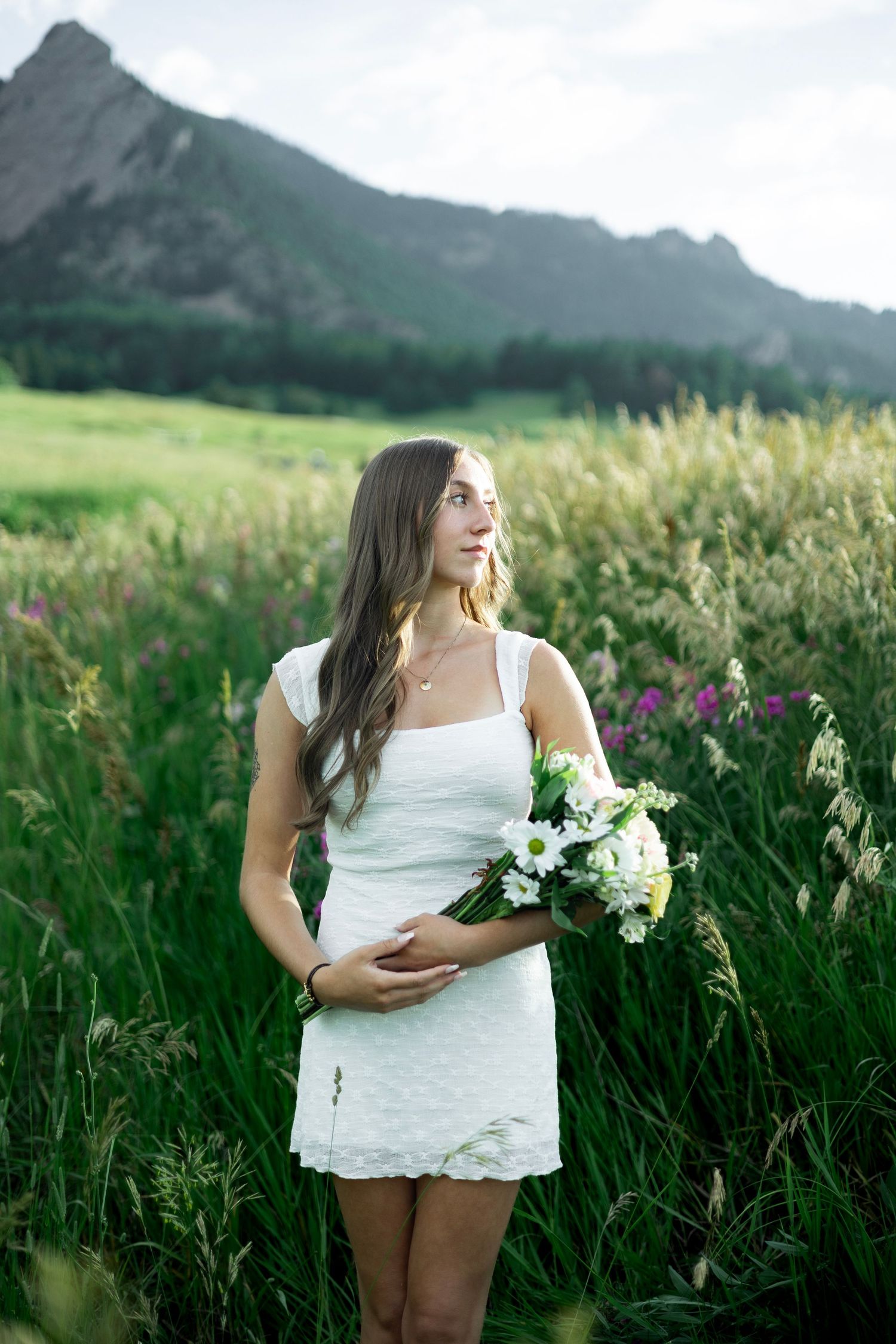 A subject in a white dress stands amid tall wildflowers and grasses with scenic mountains in the background.