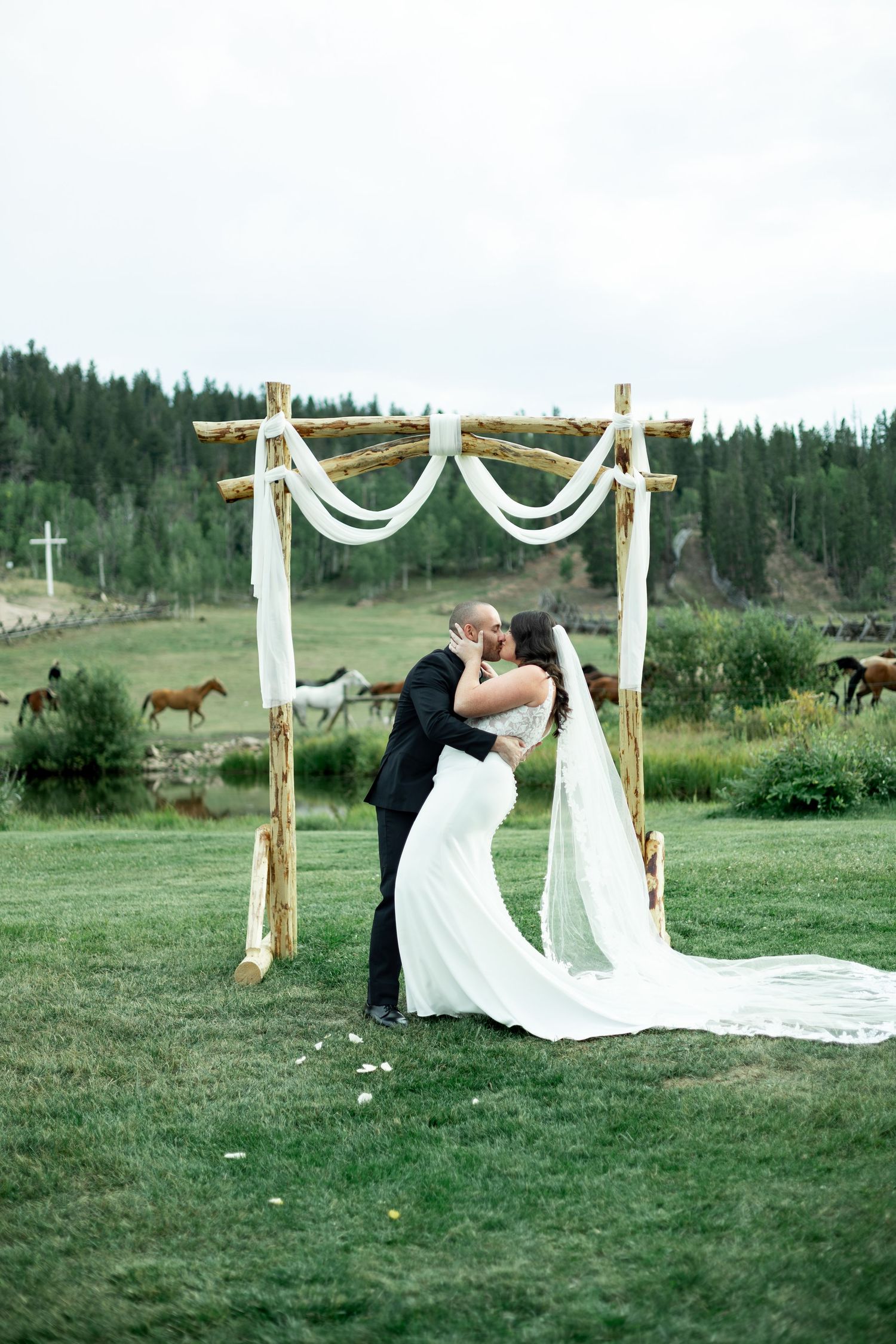 Romantic outdoor wedding ceremony with white draped arch and couple sharing a kiss in a scenic mountain meadow setting.