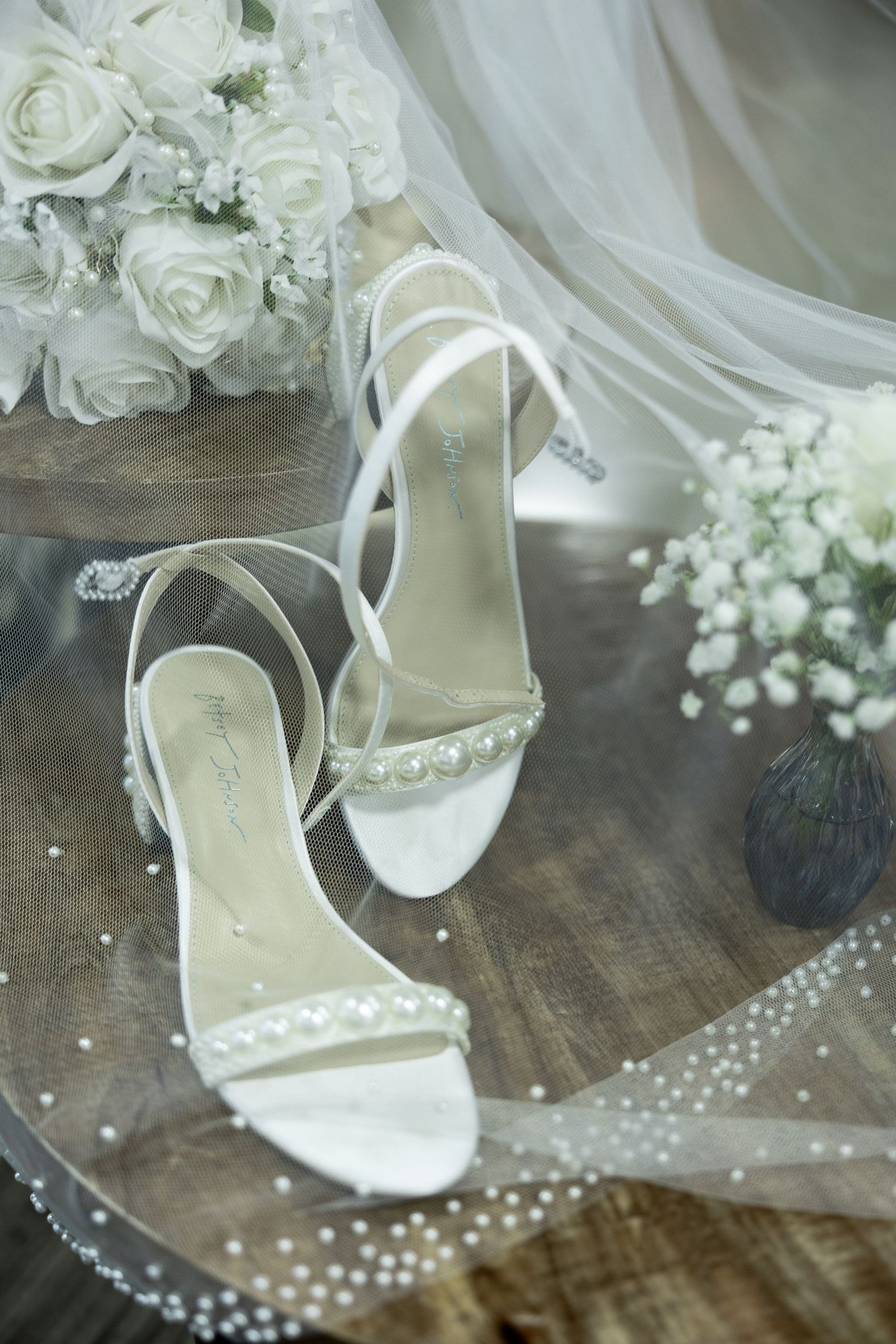 Elegant white pearl-embellished bridal heels on wooden table with rose bouquet and tulle veil.