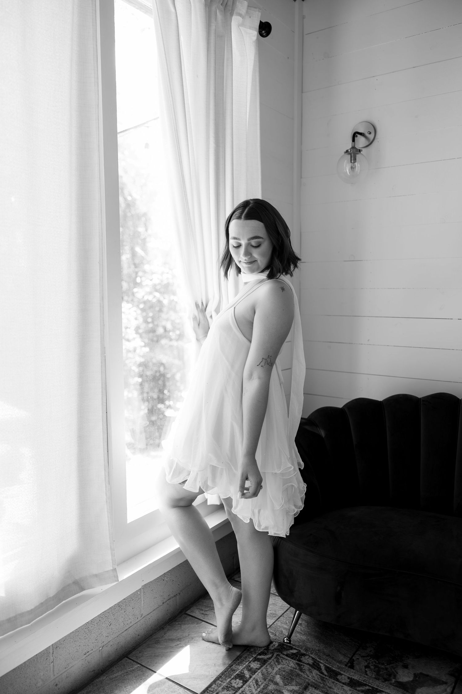 Black and white photo of barefoot woman in white chiffon dress standing by sunlit window near velvet sofa.