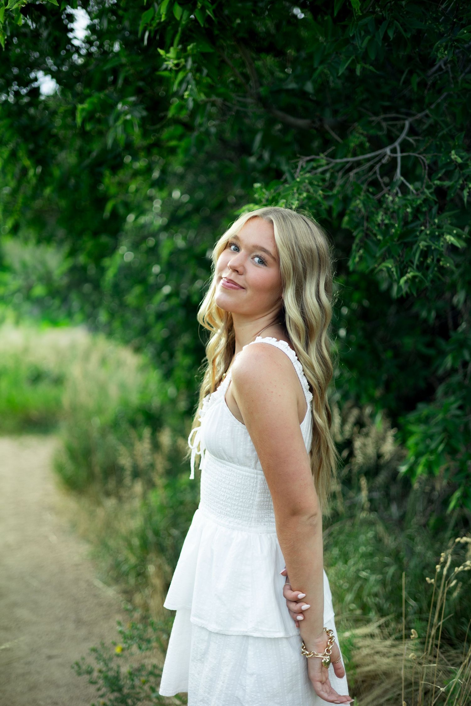 Young blonde woman in white tiered dress poses on a nature path surrounded by lush green trees.