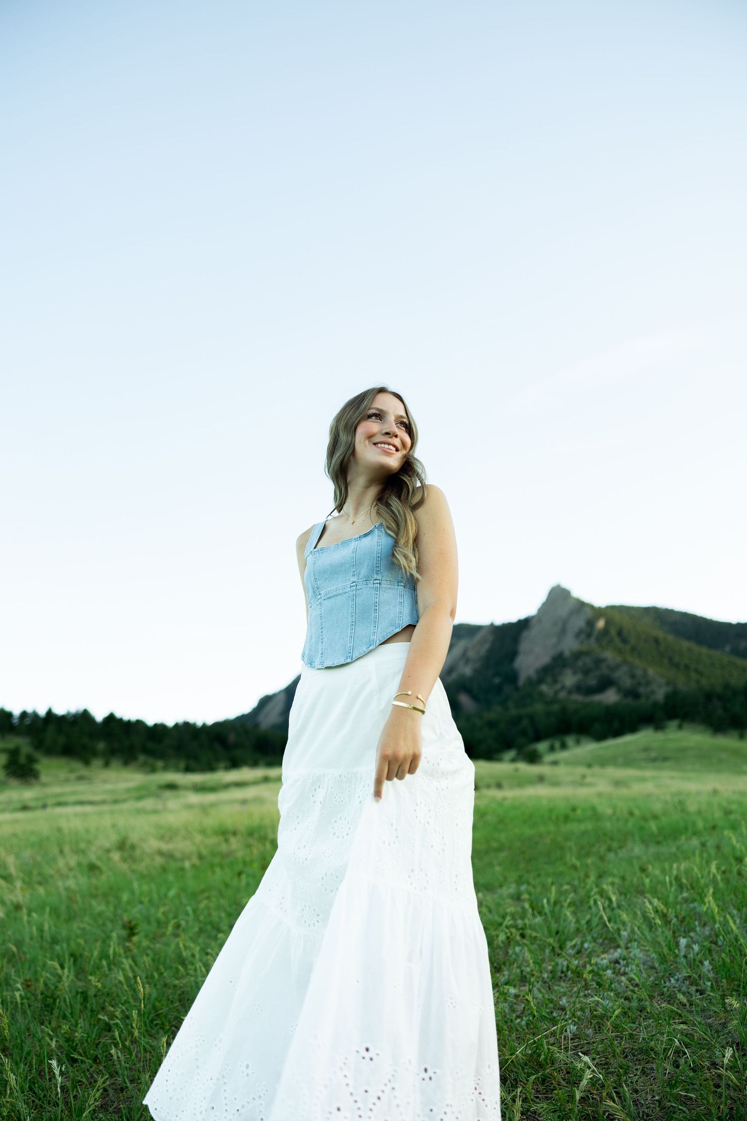 Young woman in white maxi skirt and blue top smiling in green meadow with rocky mountains in background.
