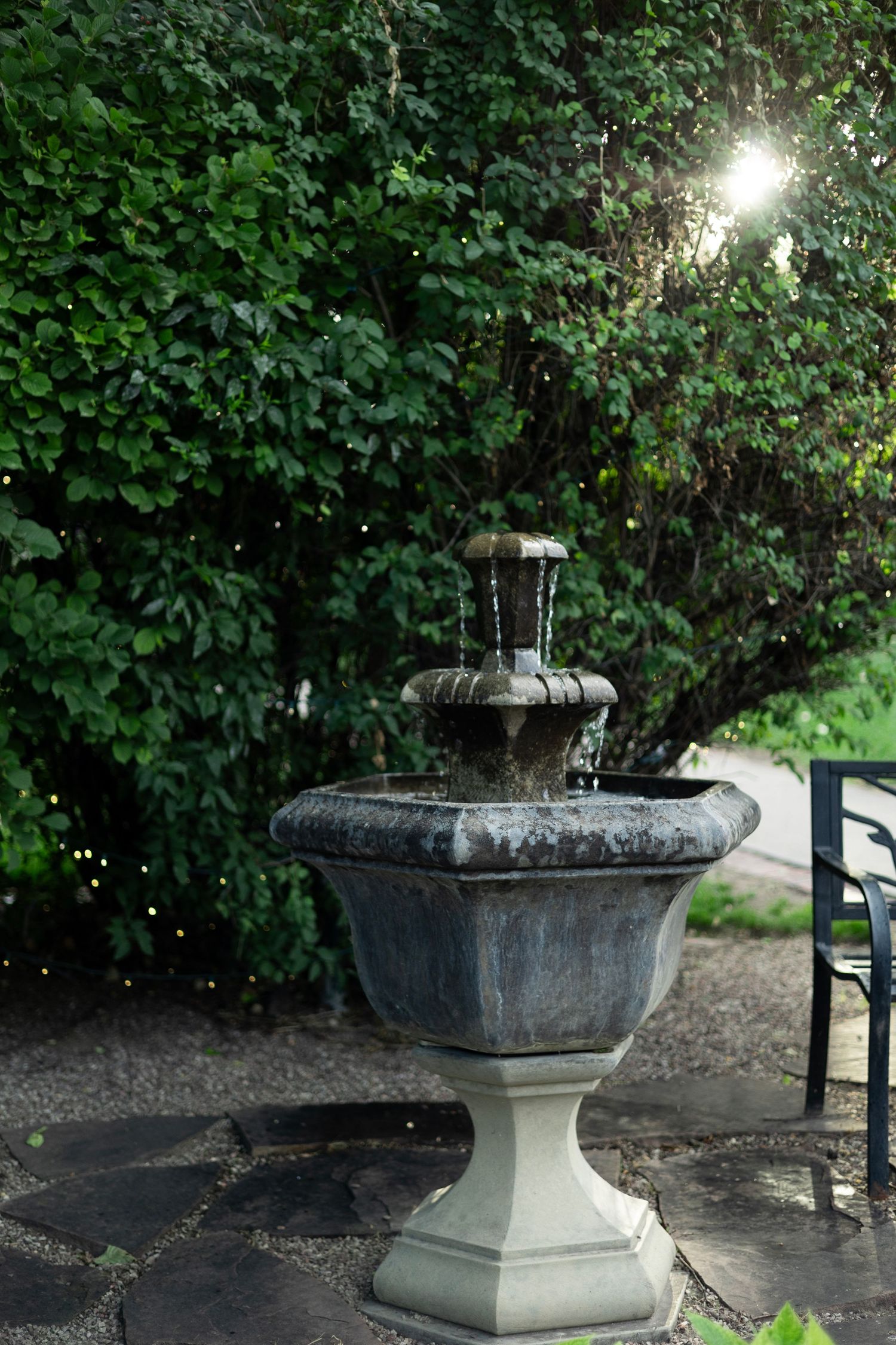 A stone fountain bubbles in a garden setting with sunlight peeking through dense foliage.