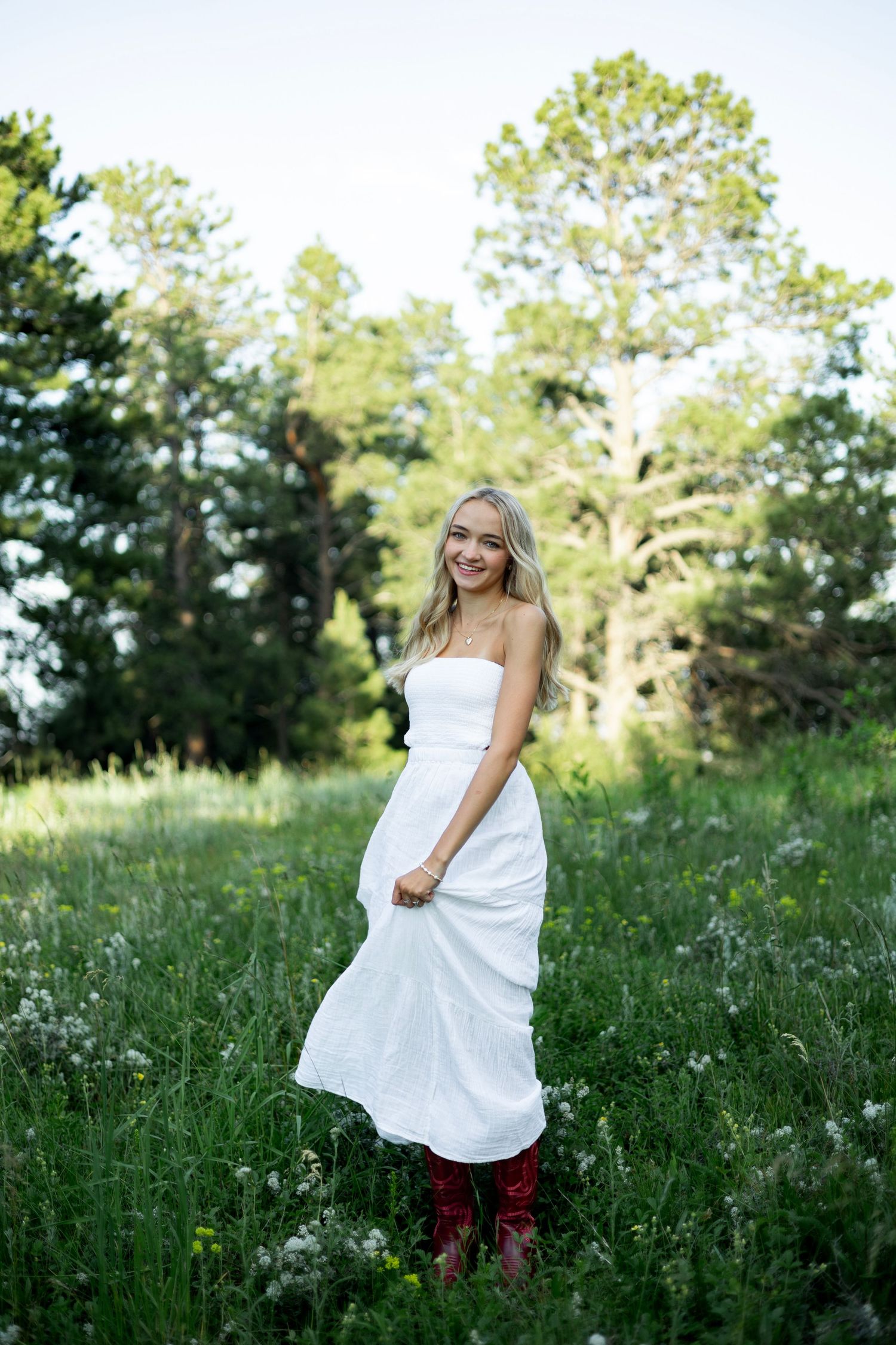 A dreamy photoshoot in a wildflower meadow shows a white dress against green grass and pine trees at golden hour.