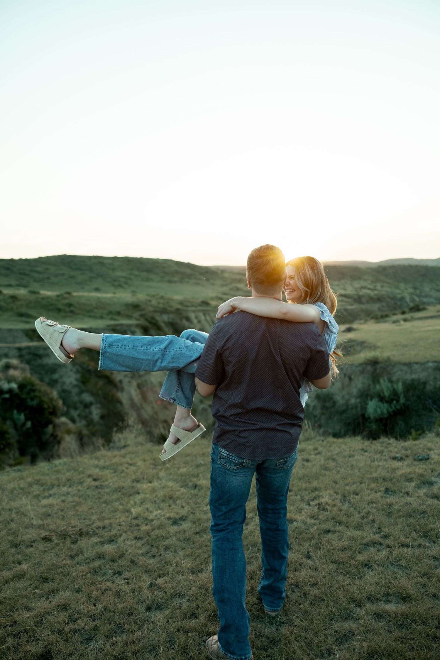 A man carries a woman in his arms at sunset overlooking green rolling hills, sharing a romantic moment outdoors.
