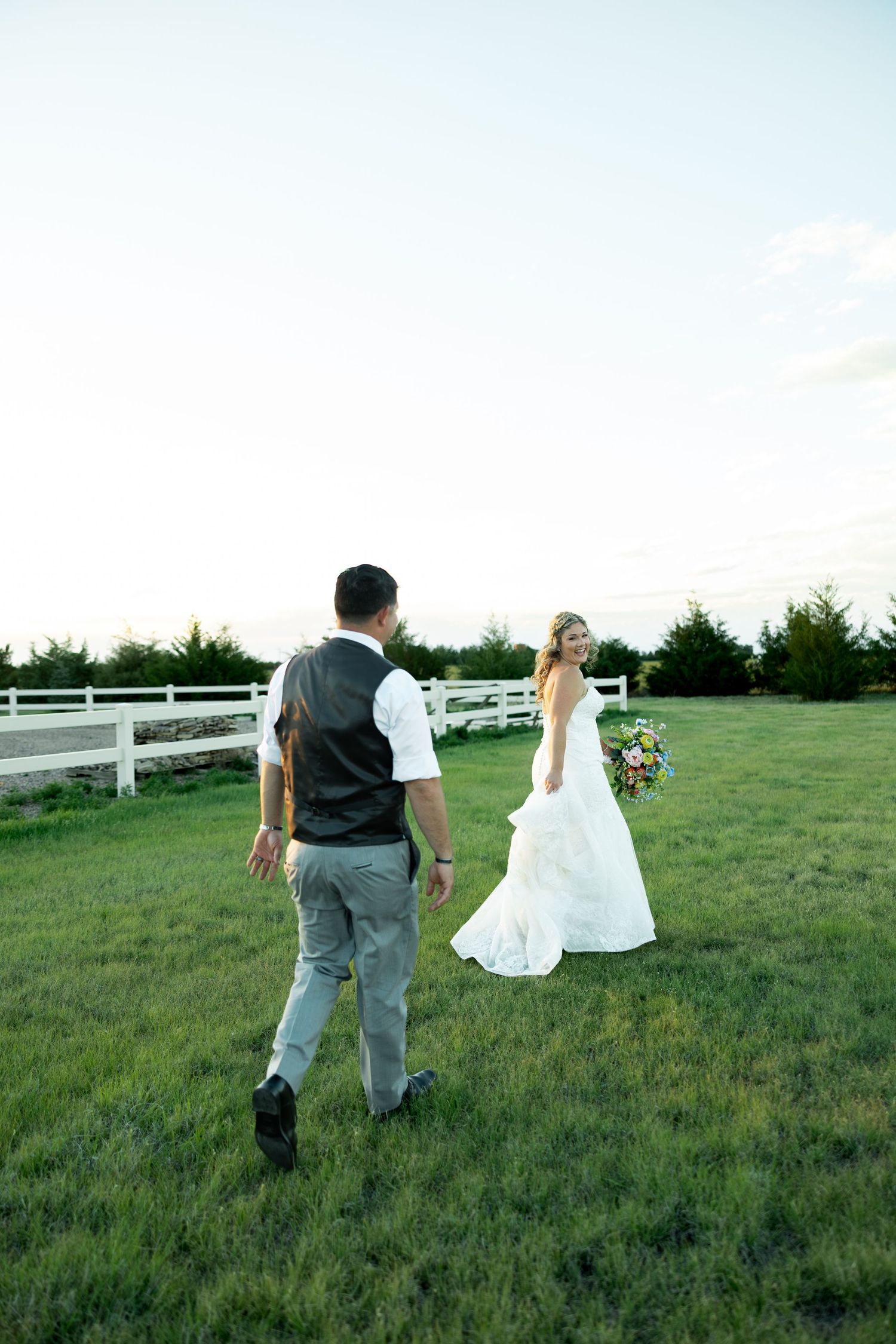 Bride in white gown holding bouquet walks with groom in vest across green lawn at outdoor wedding venue.