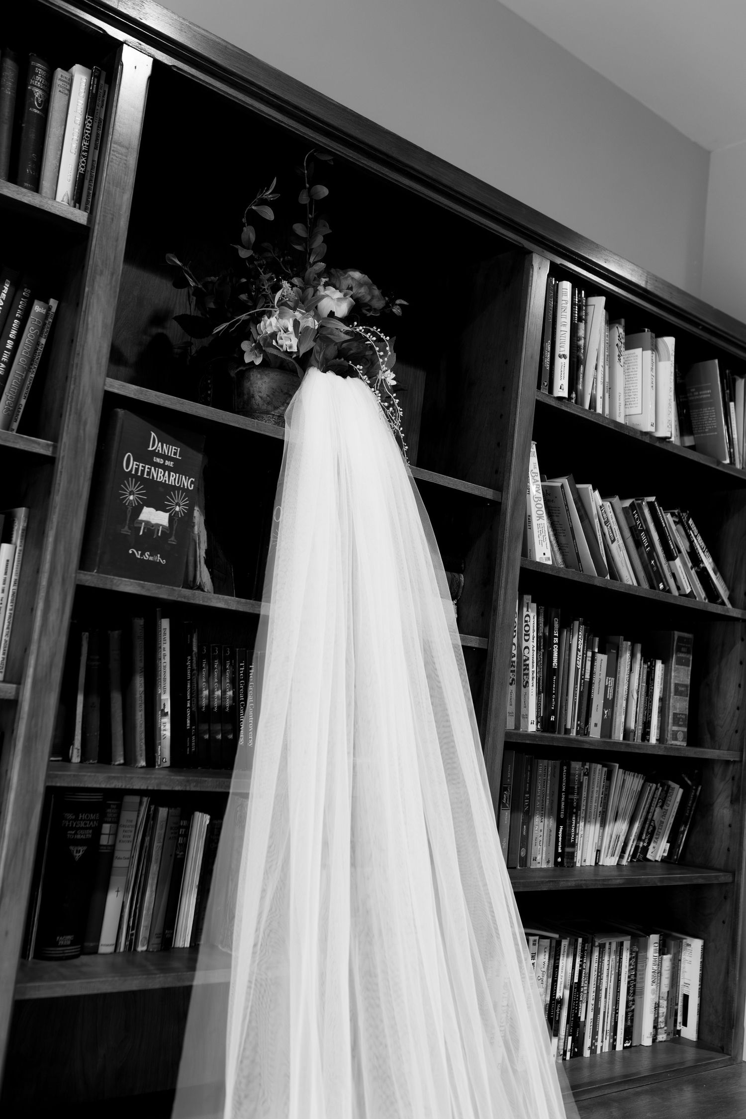 A white wedding veil hangs from a wooden bookshelf filled with books in a black and white photograph.