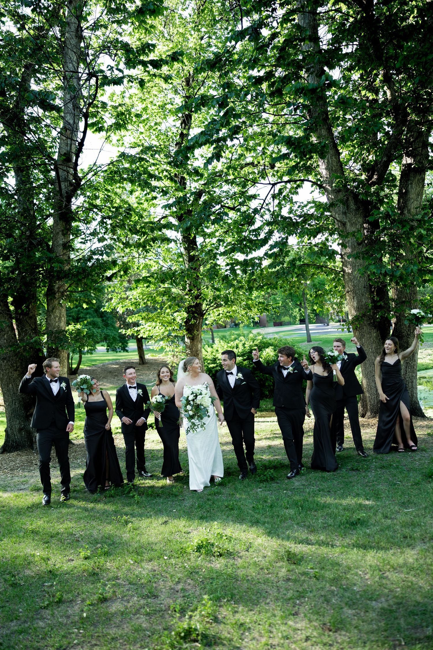 Wedding party in black attire walks with bride in white gown through a lush green park setting.