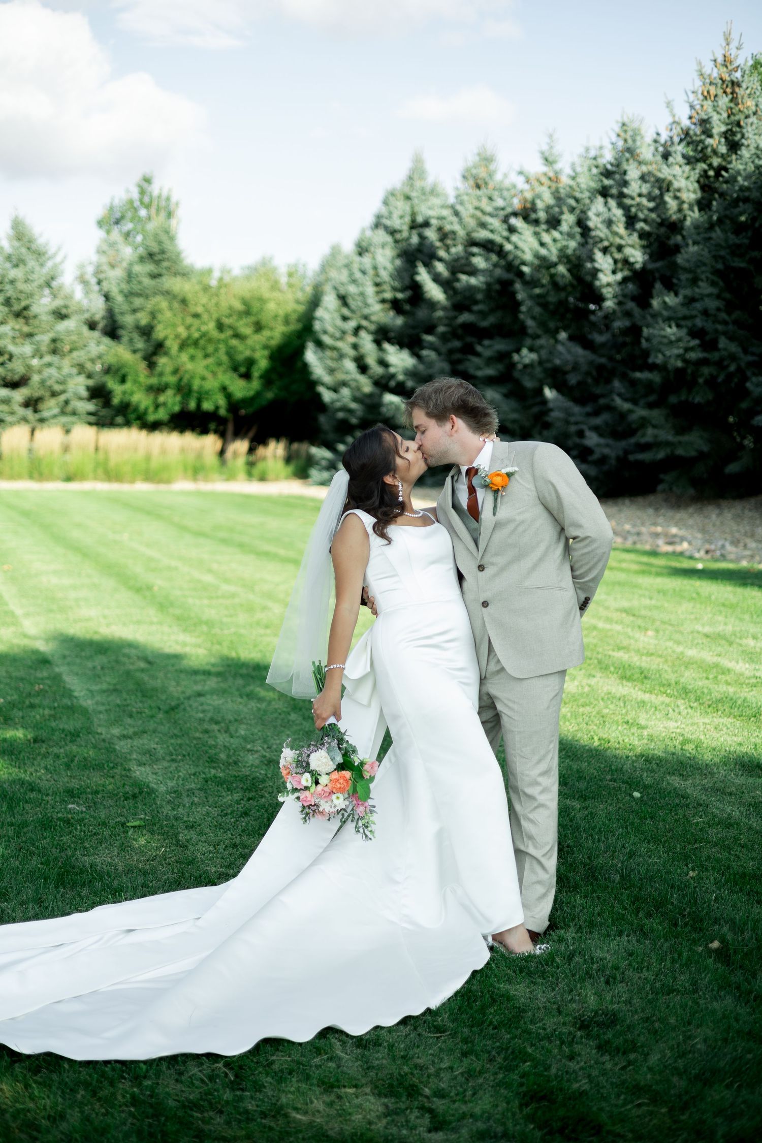 Bride and groom share a kiss on lush green lawn, she holds colorful bouquet in white gown with long train, he wears tan suit.