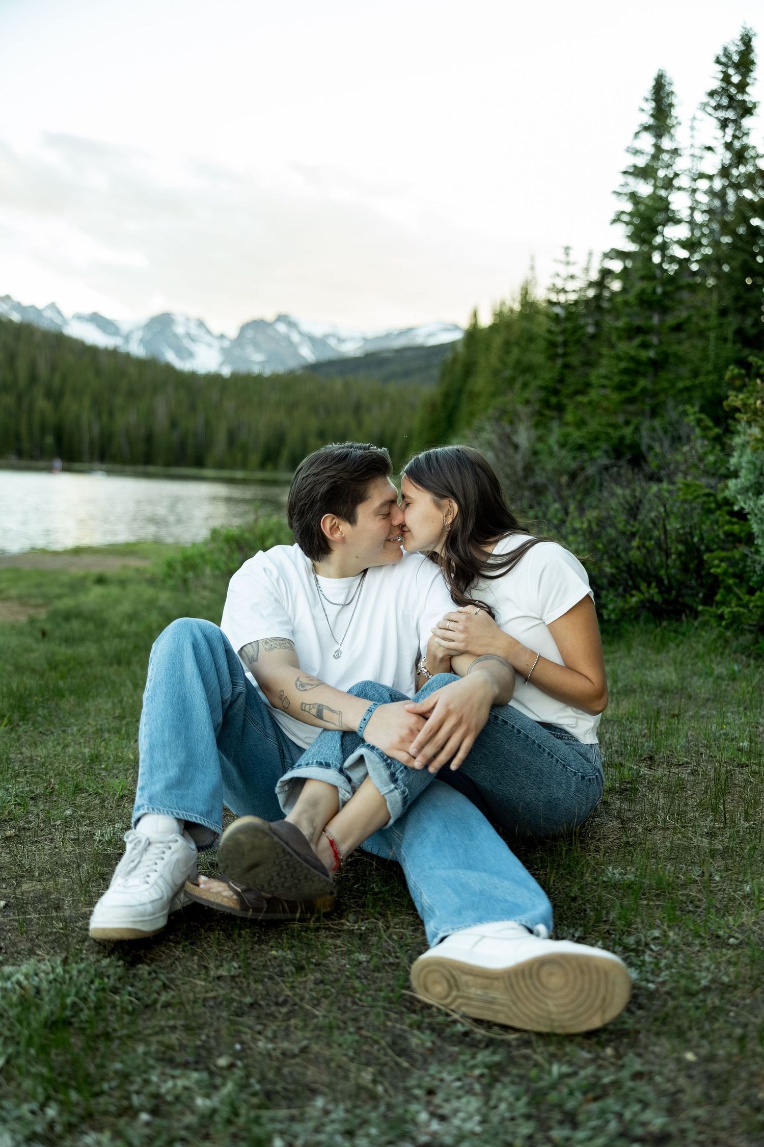 Romantic couple in white tees and jeans sitting close together near a mountain lake, sharing an intimate moment outdoors.