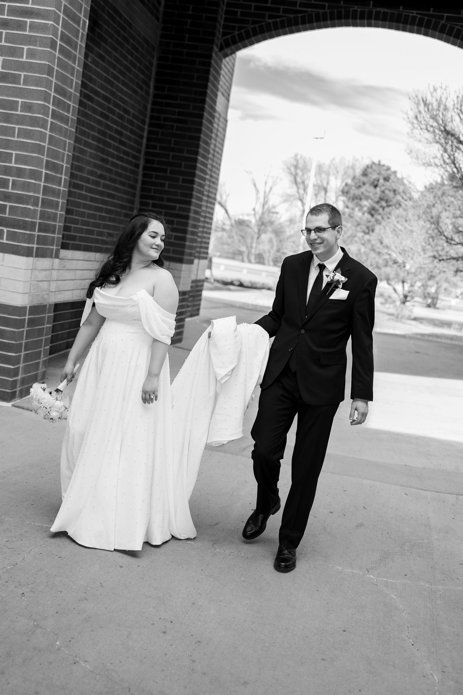 Black and white photo of a smiling bride in white gown and groom in black suit walking under brick arch.