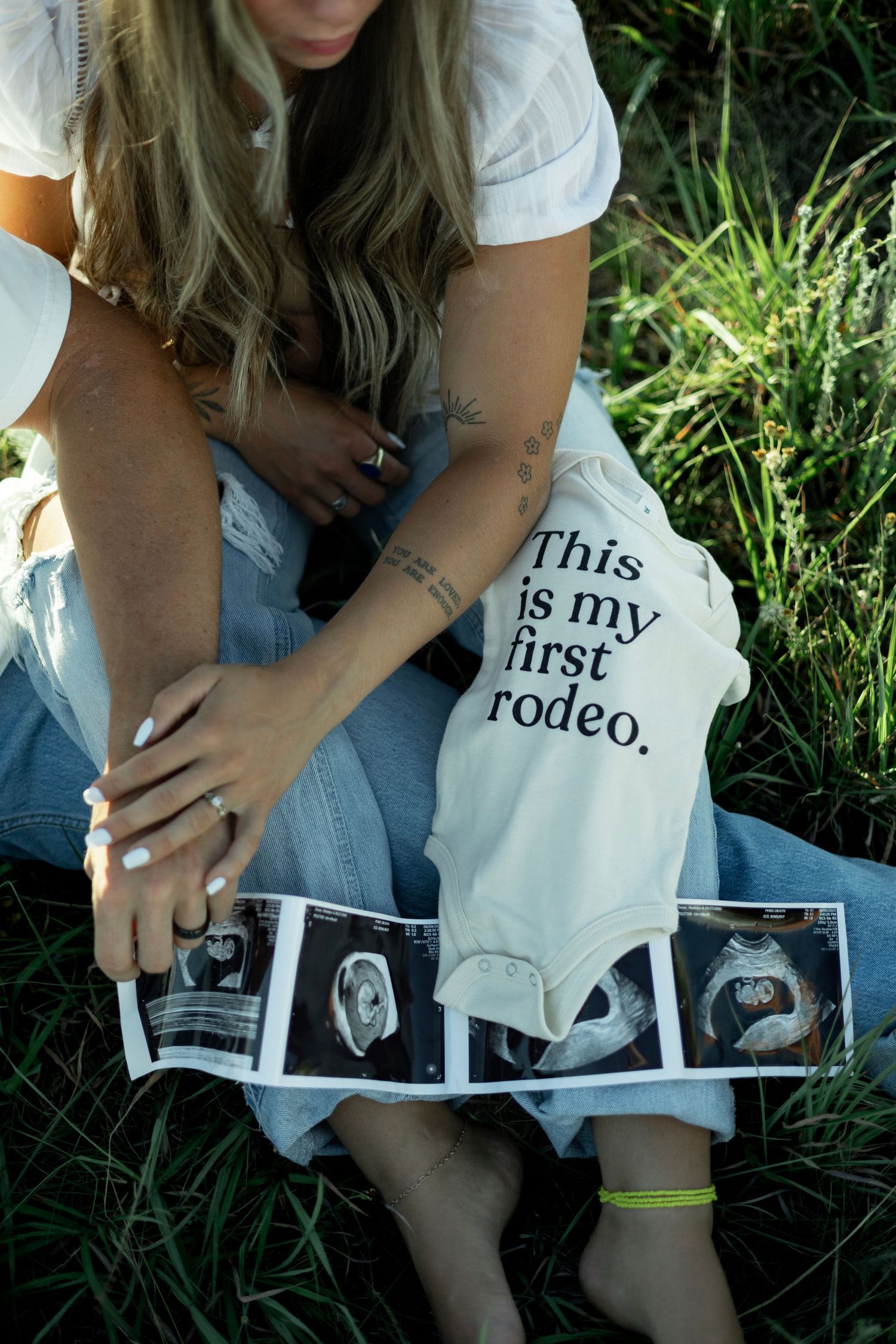 Woman in jeans sits on grass holding ultrasound photos next to a onesie that reads 'This is my first rodeo'.