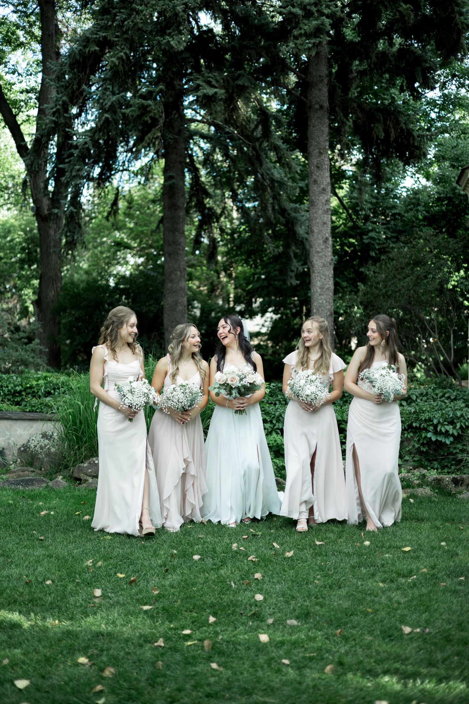 Bridesmaids in cream-colored dresses gather on grass under trees for wedding photos.
