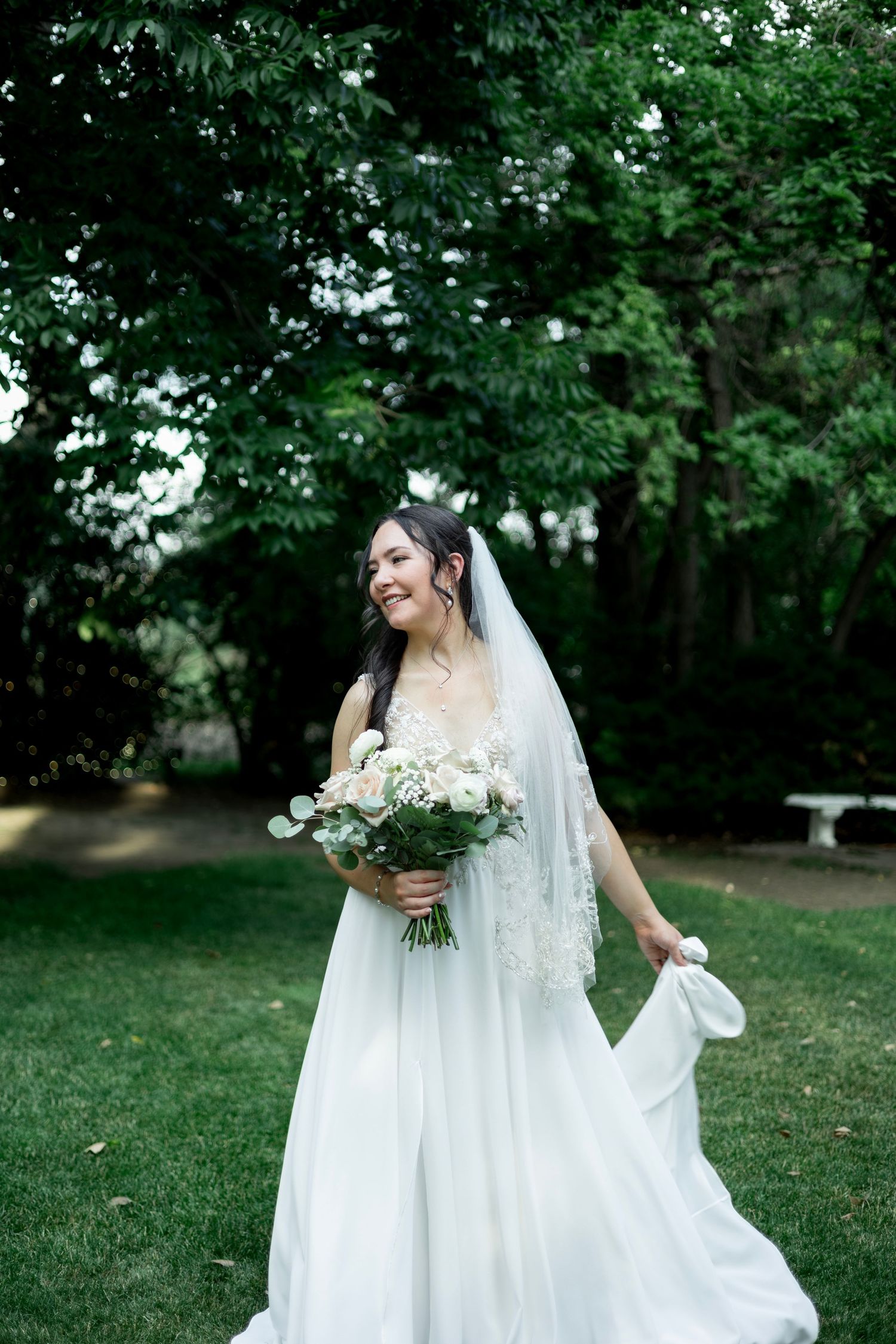 A white wedding gown and veil photographed against a lush garden backdrop with green trees.