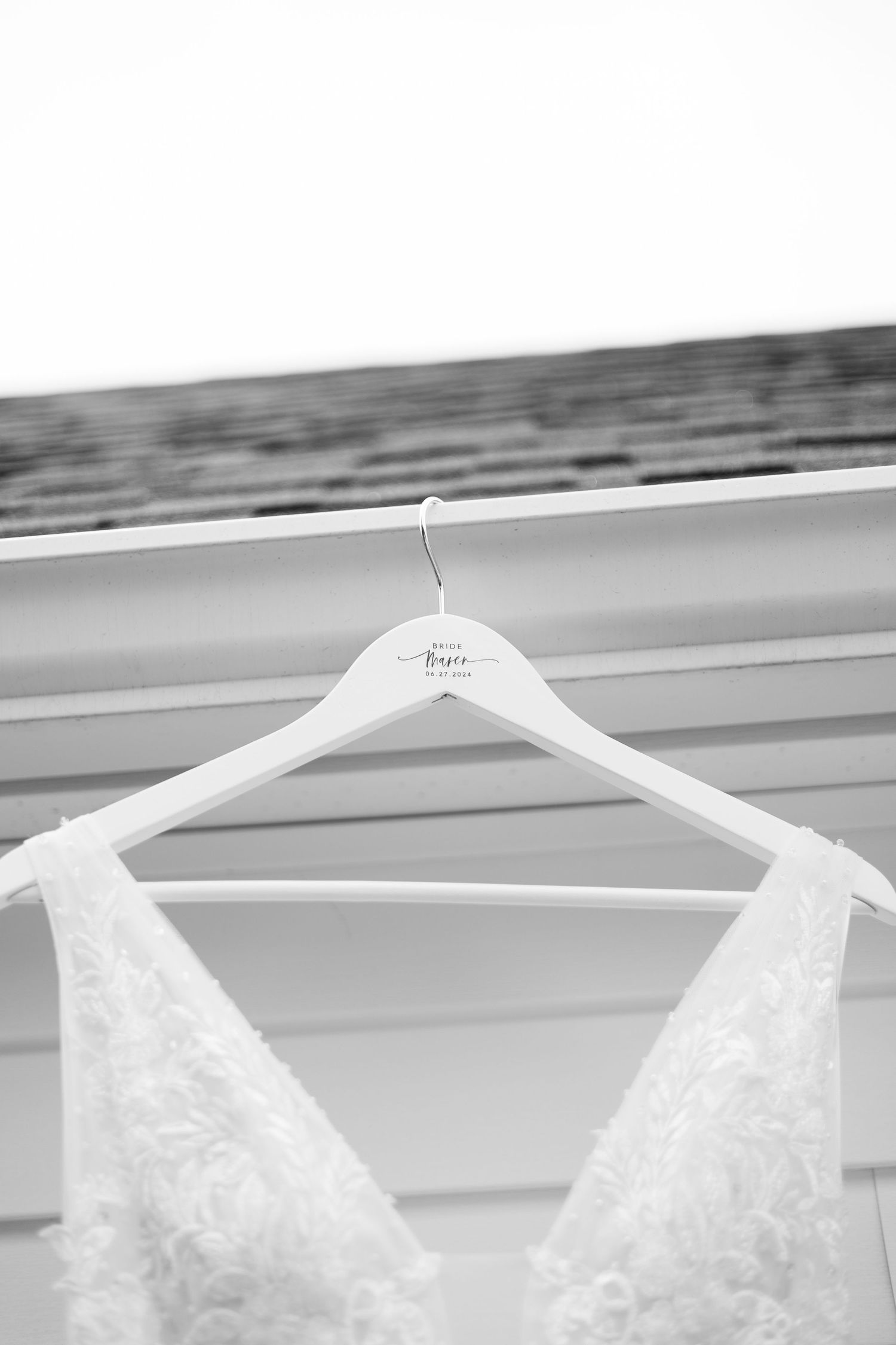 A delicate white lace wedding dress hangs on a white hanger against a light background.