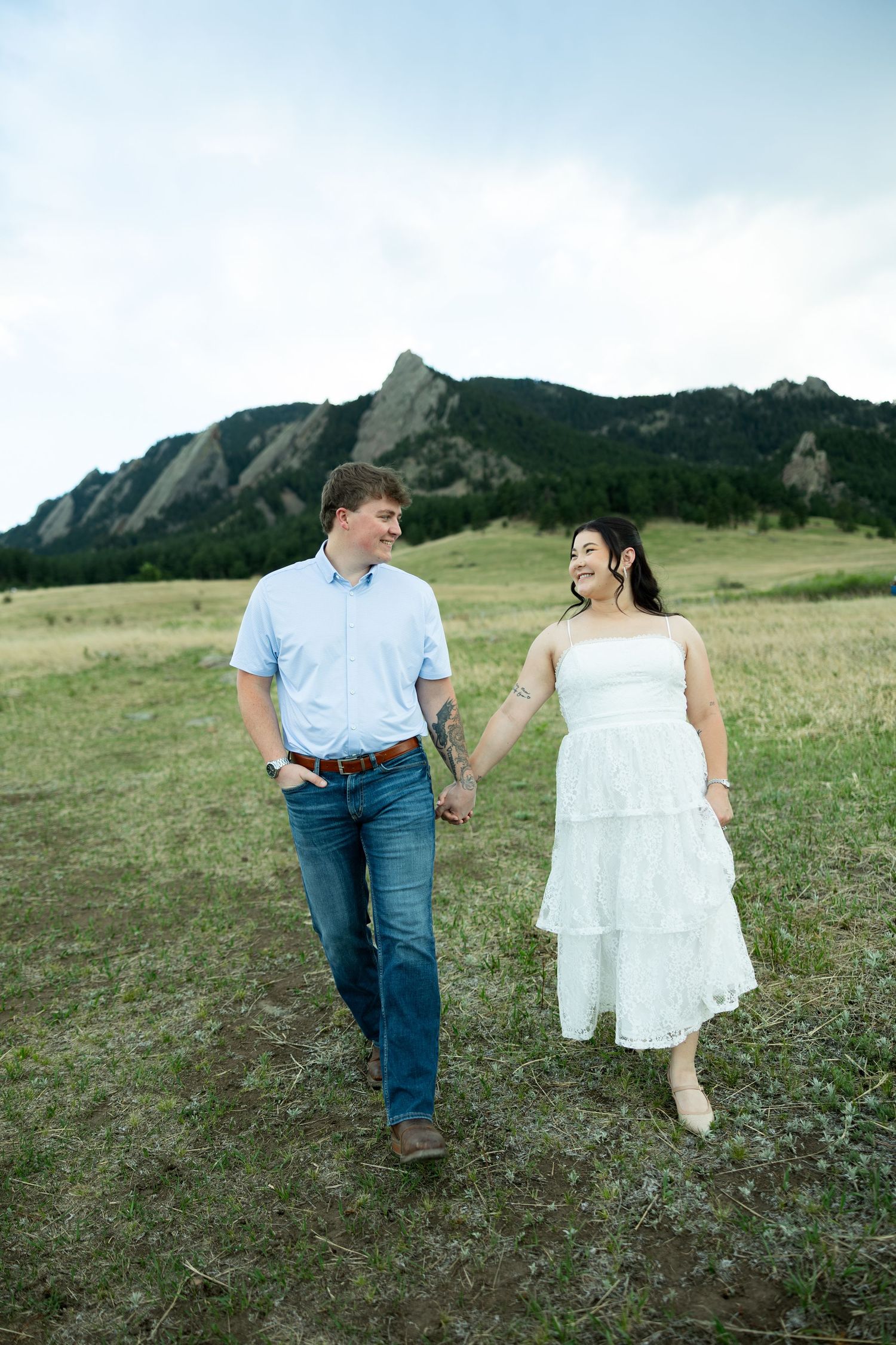 A couple holds hands walking through a green meadow with the Flatirons rock formations in Boulder, Colorado behind them.