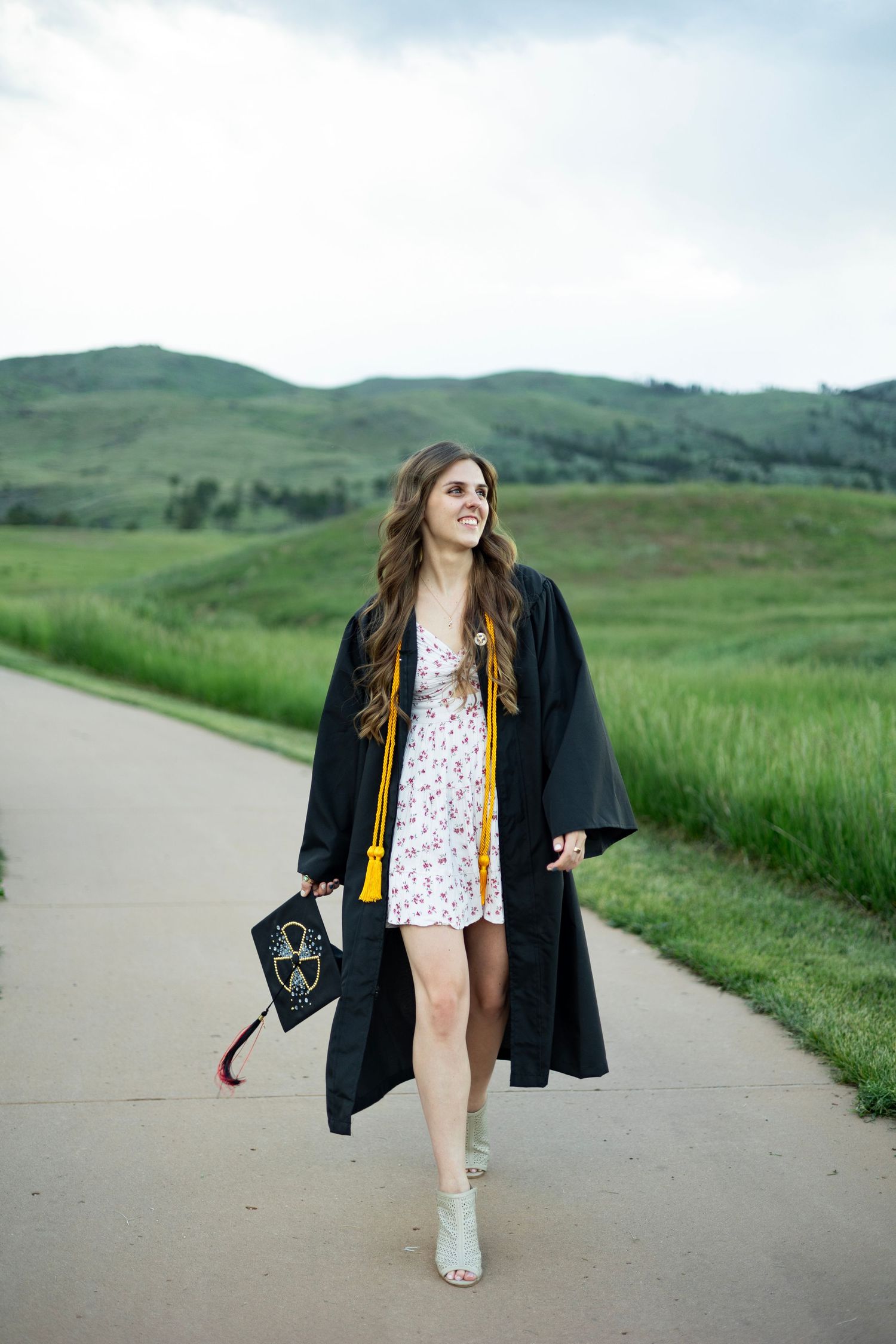 A young woman in a black graduation gown and yellow tassel walks on a path with green hills in the background.