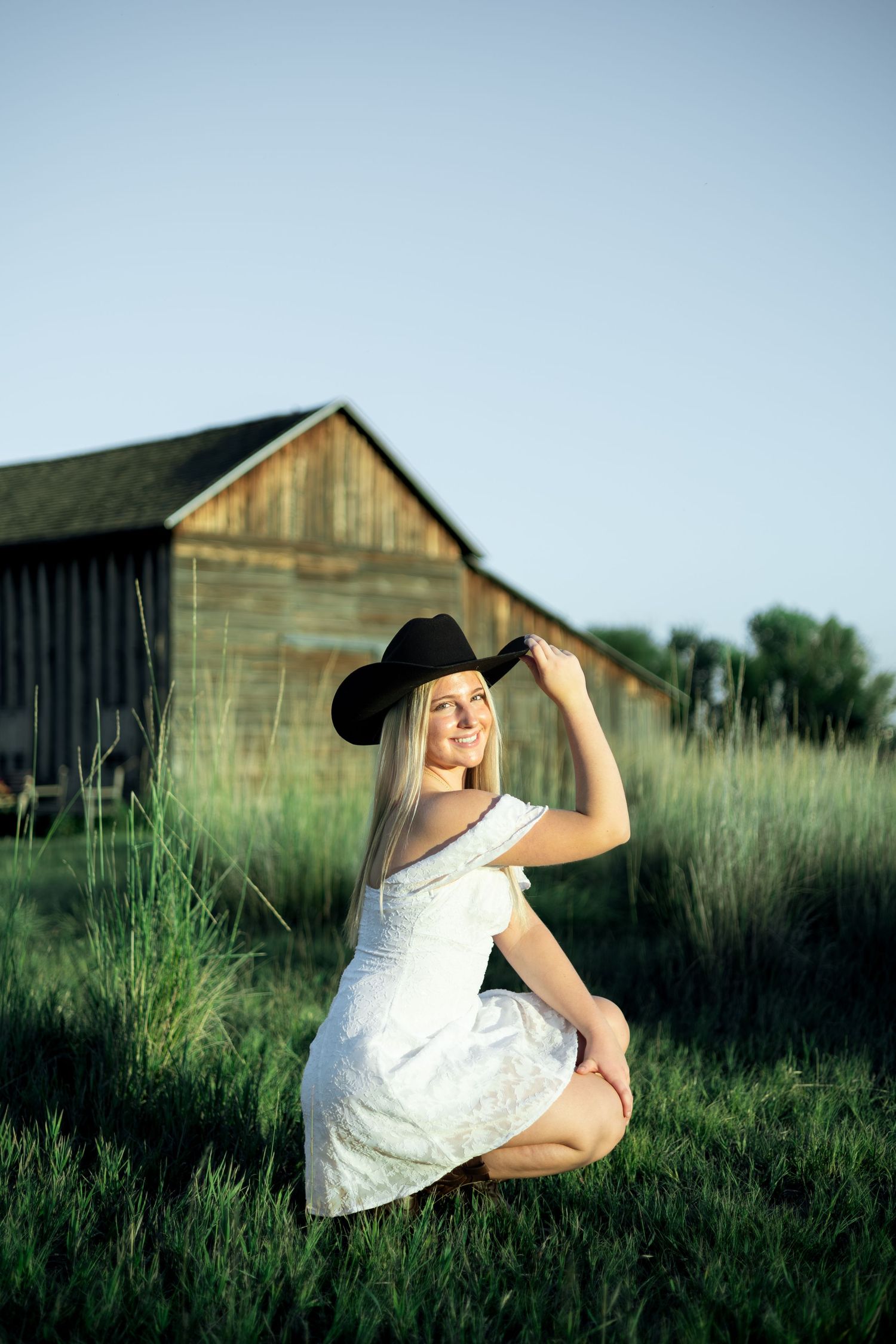 Blonde woman in white dress and black cowboy hat sitting in green grass near rustic wooden barn at sunset.