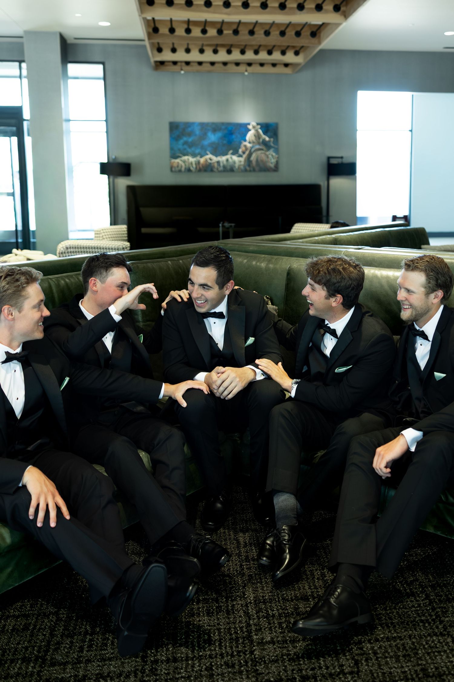Five groomsmen in black tuxedos laughing and relaxing together on a green sofa in an elegant venue.