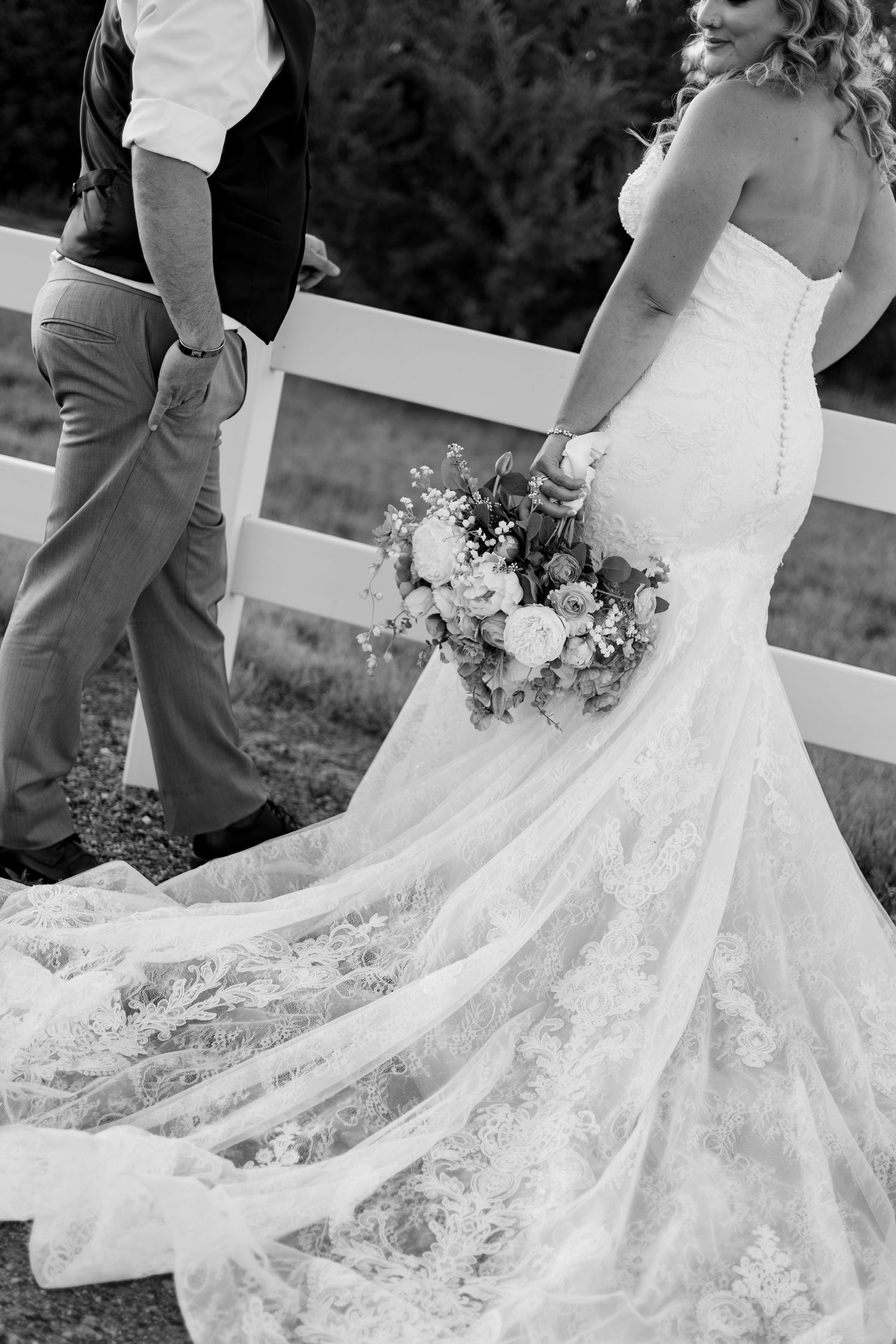 Black and white photo of bride in lace mermaid gown holding floral bouquet walking with groom near white fence.