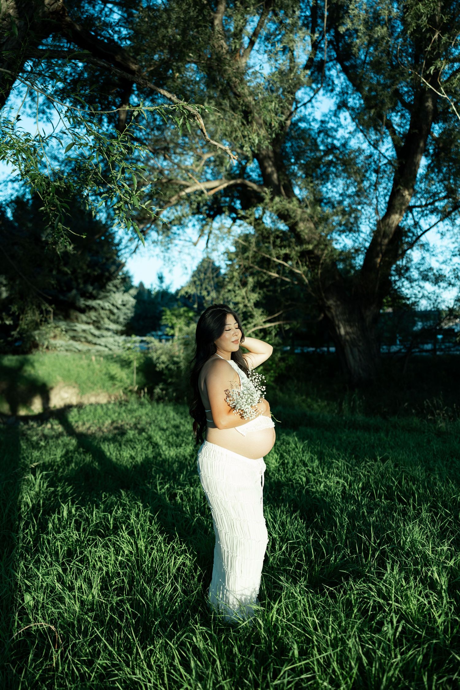 Pregnant woman in white skirt and floral top stands in lush green grass under large trees in golden hour light.