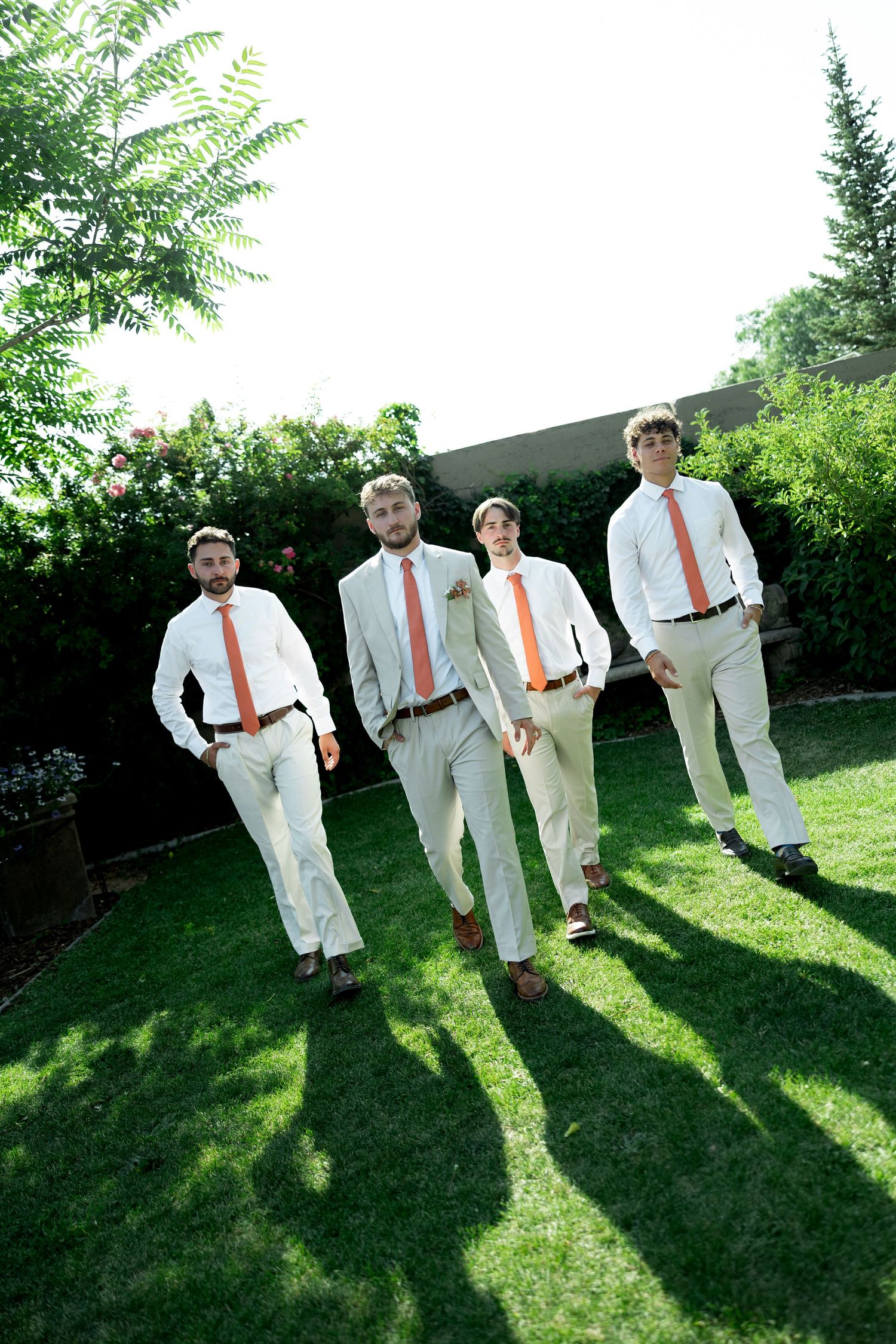 Four groomsmen in white shirts and orange ties walk across green lawn during outdoor wedding celebration.