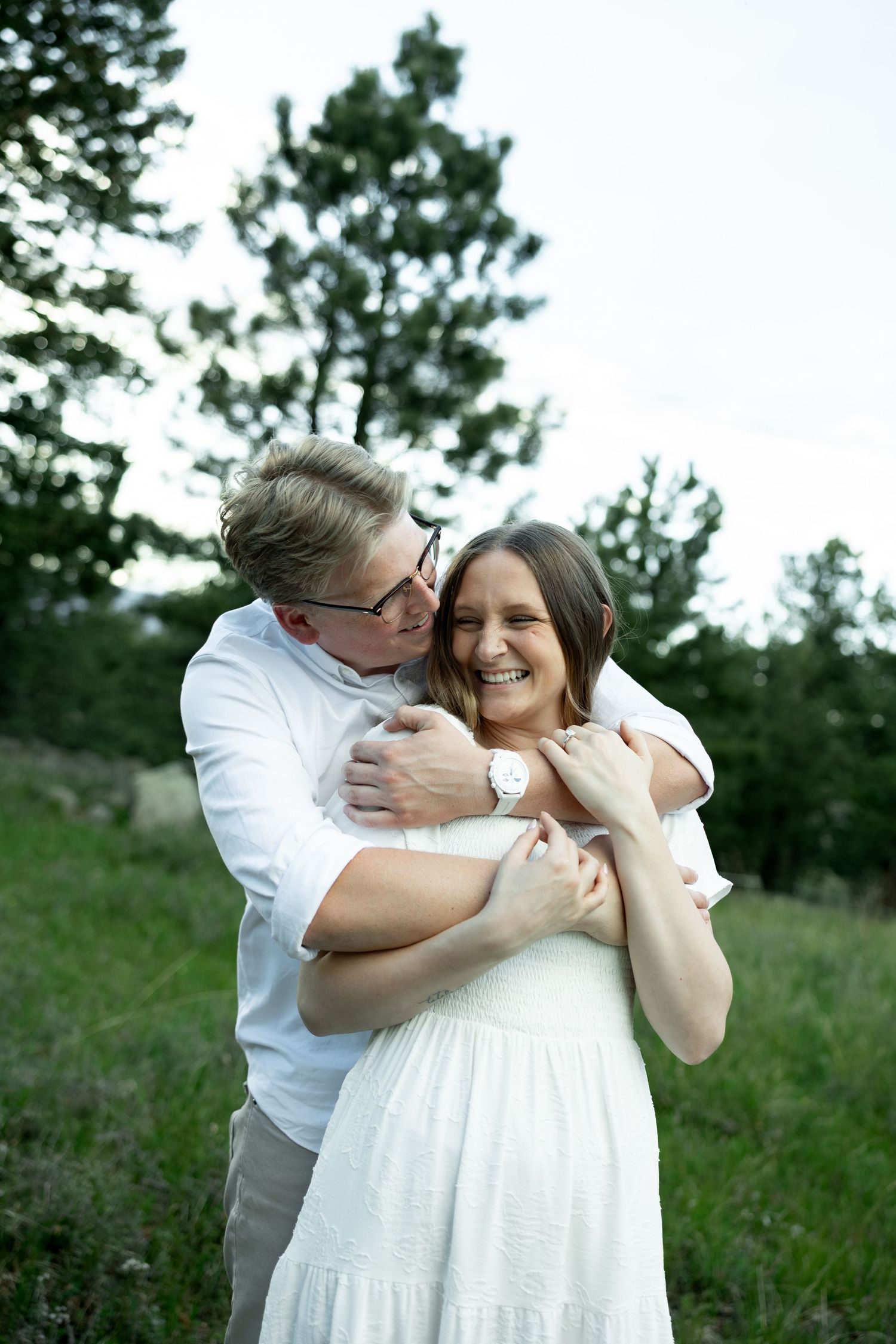 Man in white shirt hugging smiling woman in white dress outdoors in a green field.