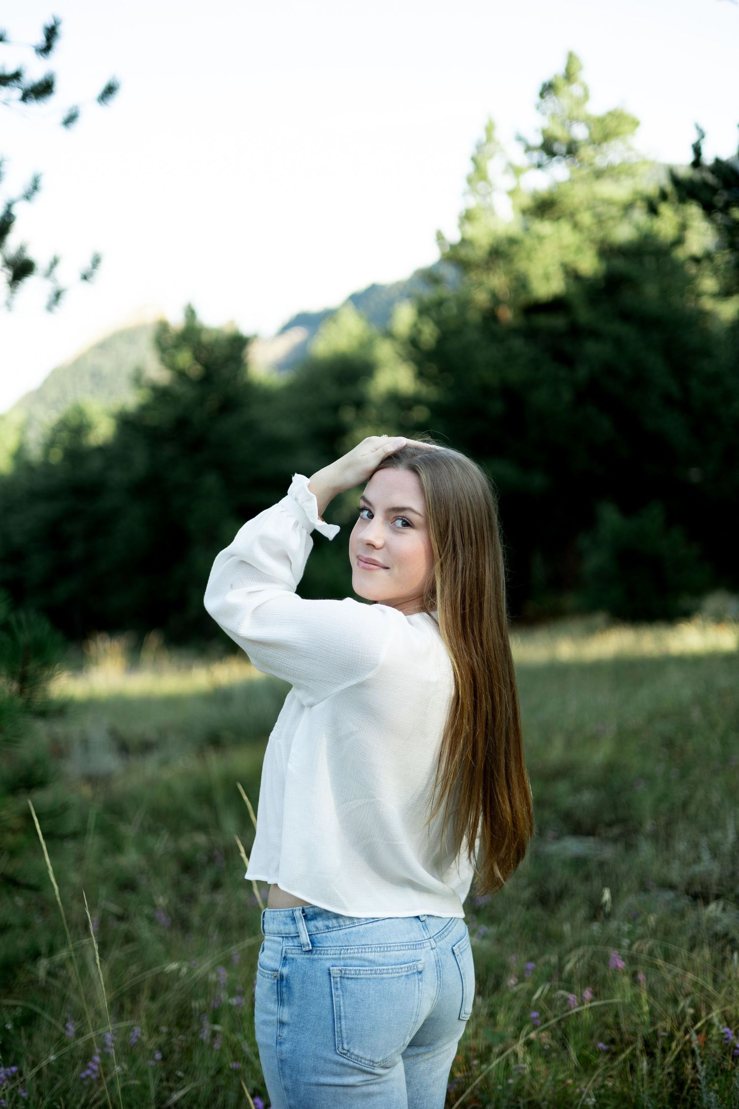 A person in a white sweater and jeans poses thoughtfully in a grassy field with mountains in the background.