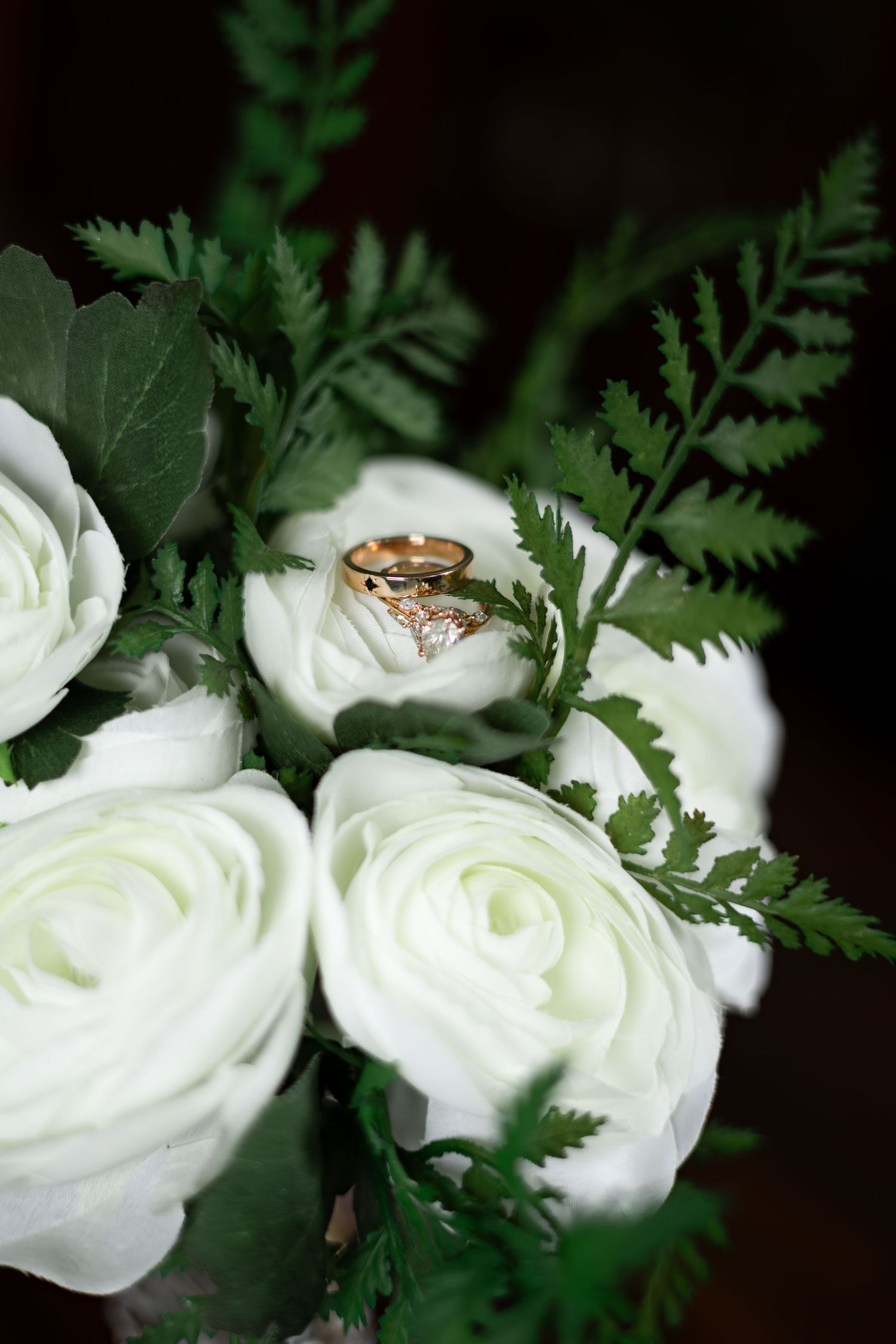 Two golden wedding rings nestled among delicate white ranunculus flowers and fern leaves.