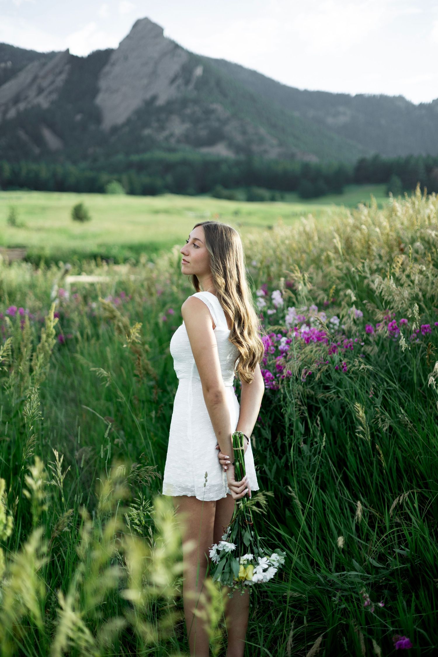 A figure in a white dress stands in a wildflower meadow with dramatic mountain peaks rising in the background.