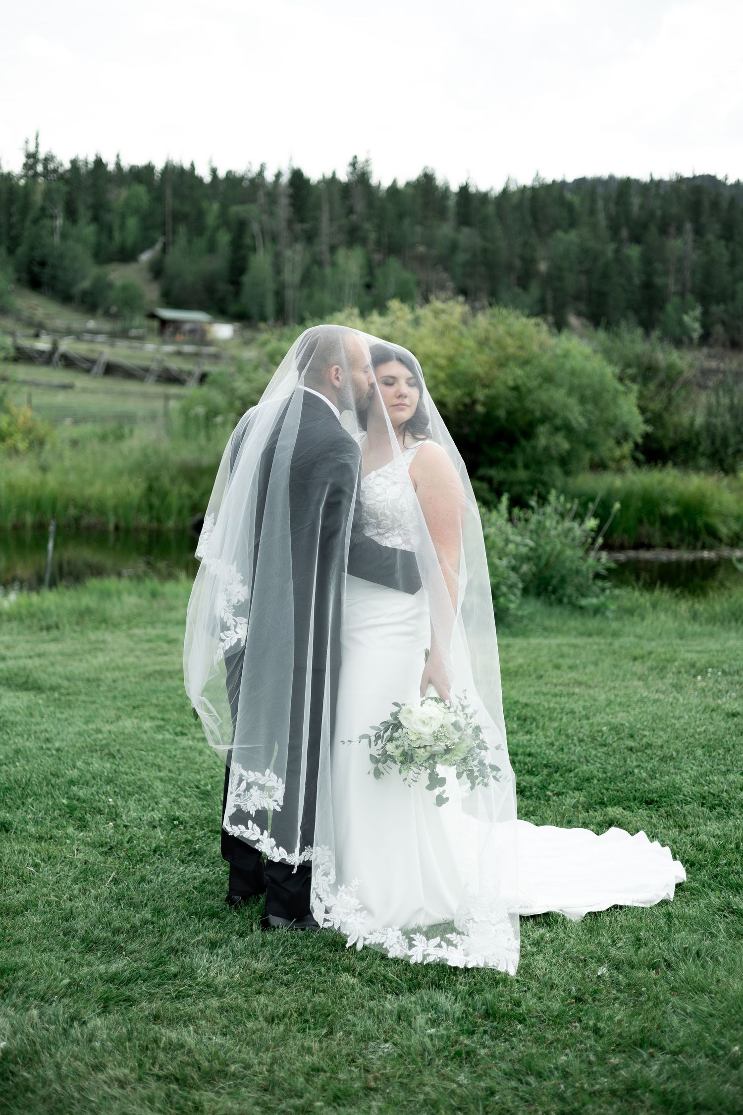 Bride and groom share a romantic kiss under a lace veil on green grass with a forest backdrop on their wedding day.