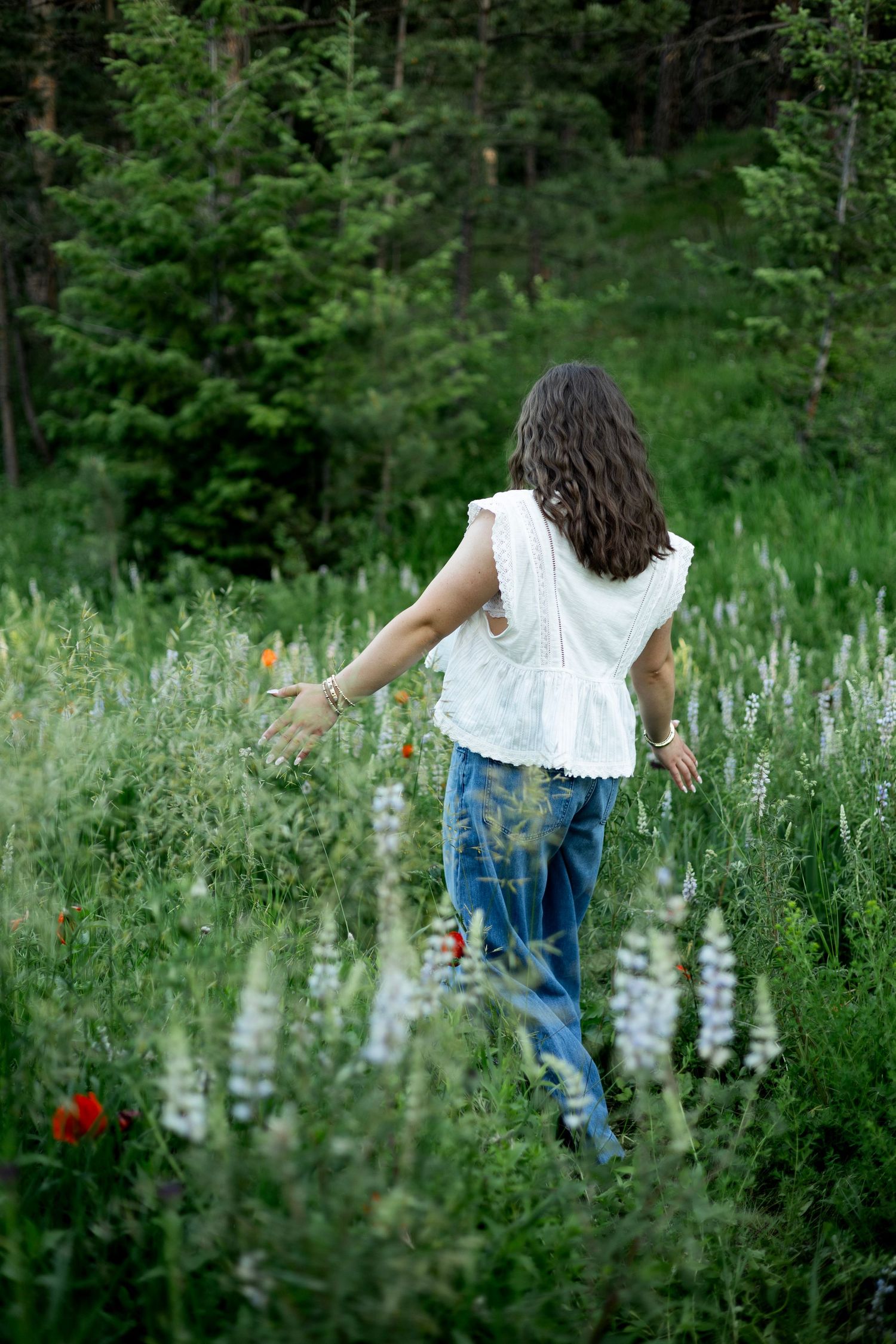 A woman in a white blouse walks through a wildflower meadow with tall lupines and red blooms in a forest setting.