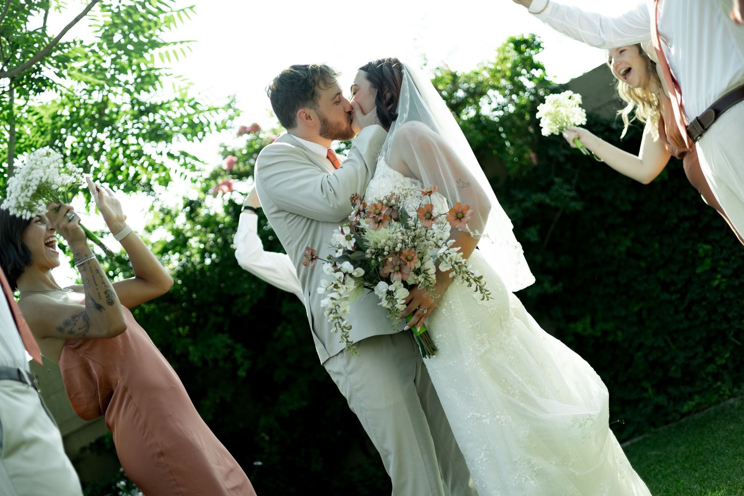 Bride and groom share first kiss at outdoor wedding ceremony surrounded by cheering bridesmaids holding bouquets.