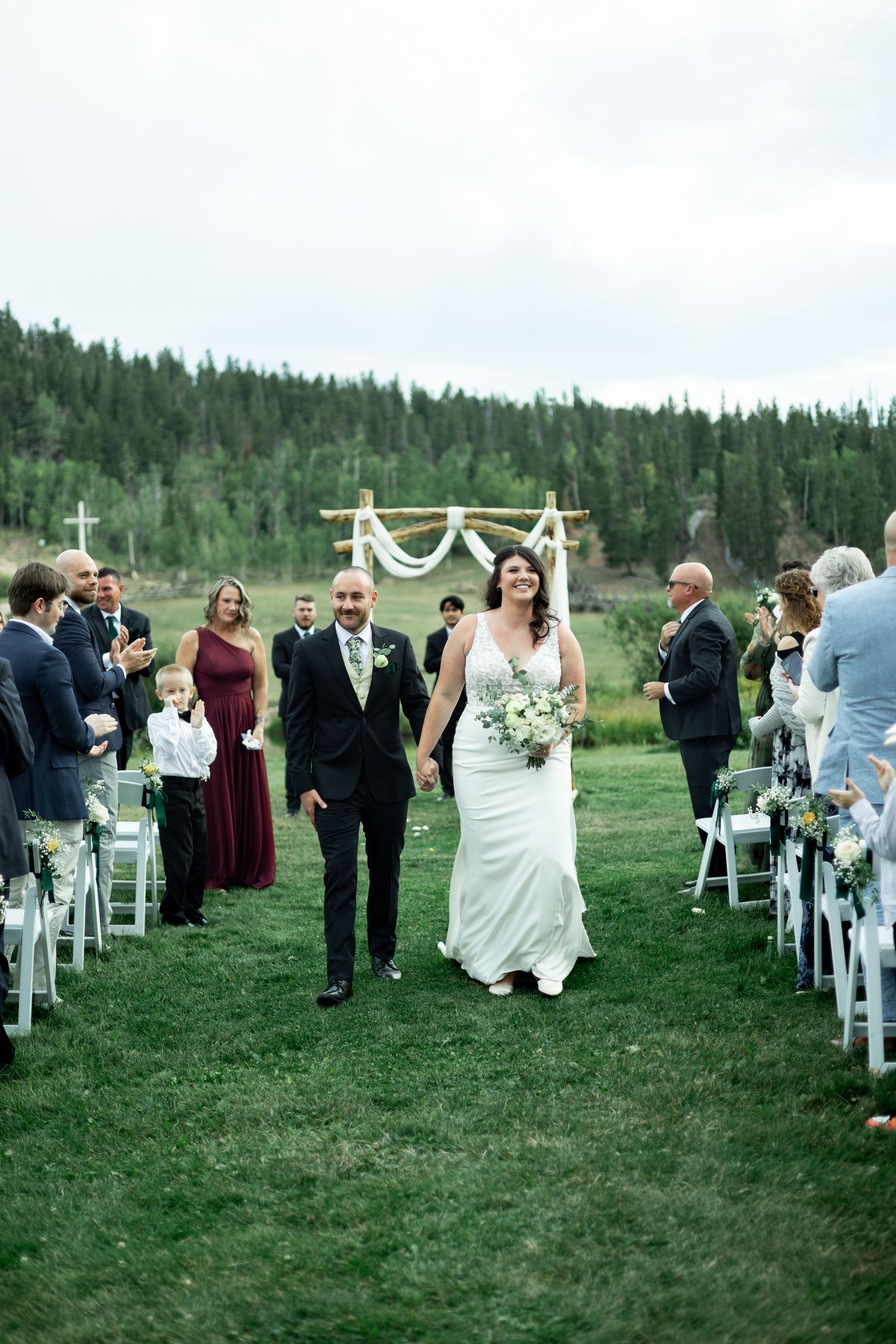 Wedding guests gather on white chairs outdoors for a rustic mountain ceremony with wooden arch and white draping.