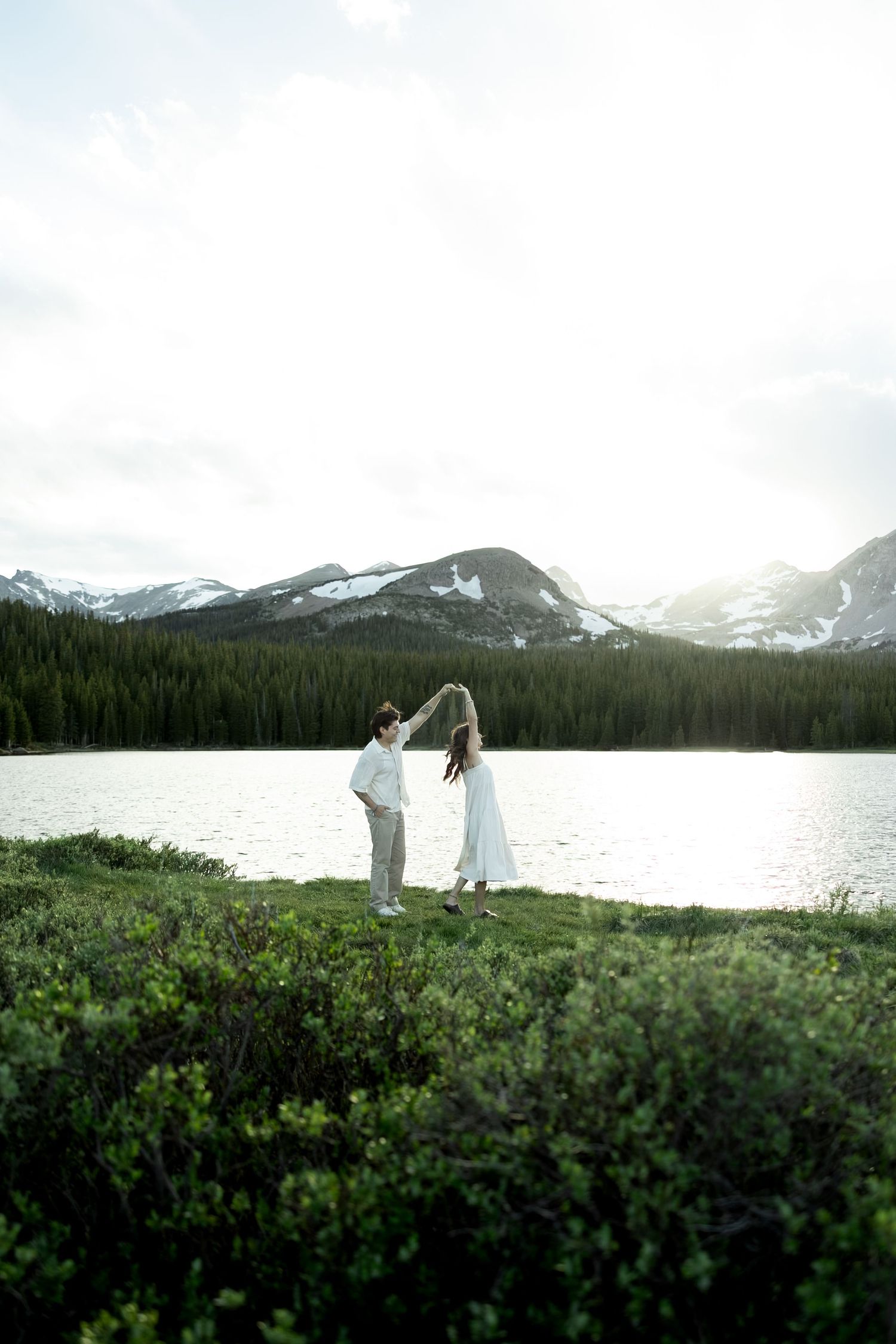Couple dancing by a mountain lake with snow-capped peaks and pine trees in the background.