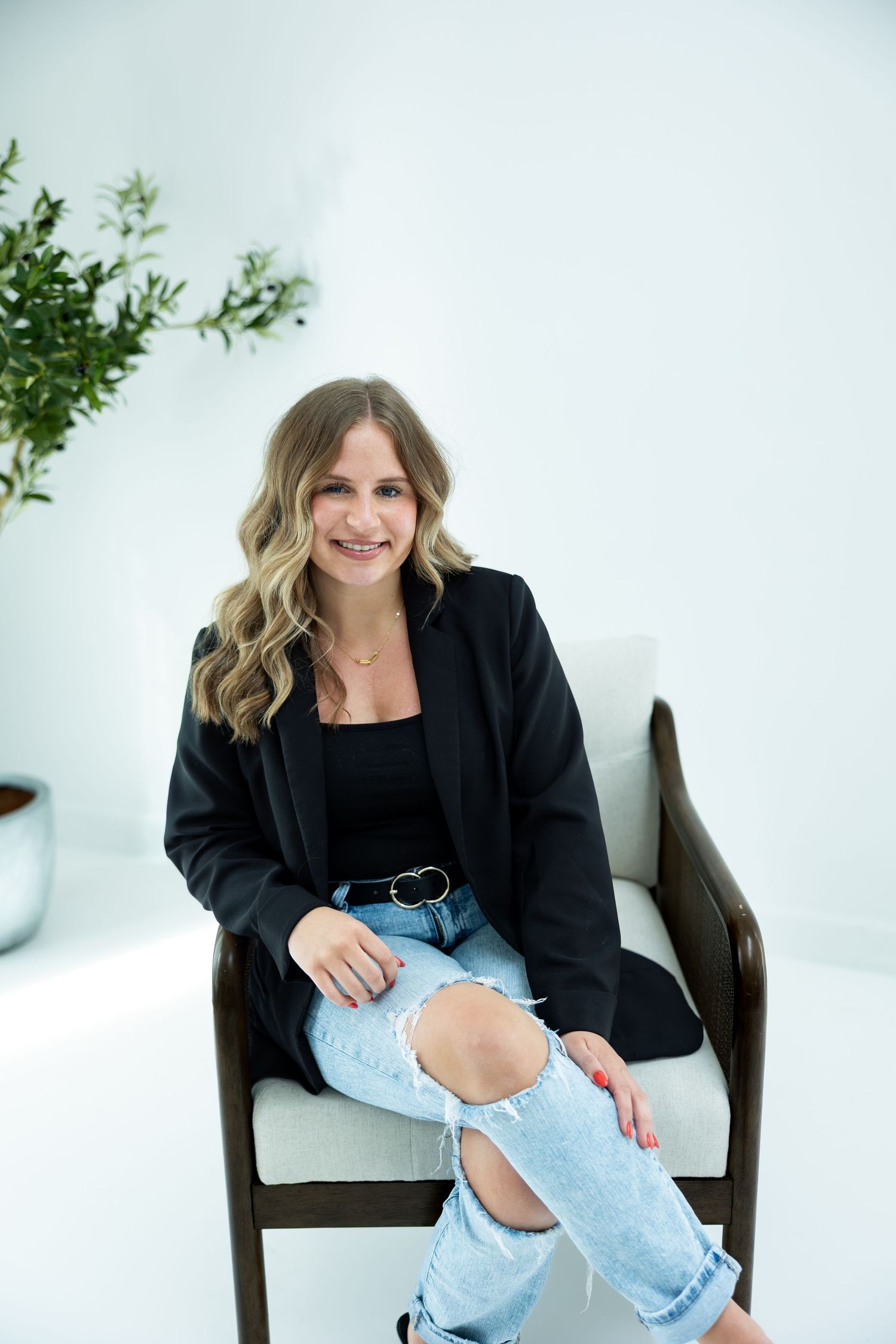 Woman in black blazer and ripped jeans sitting in a chair, smiling confidently in a bright white studio.