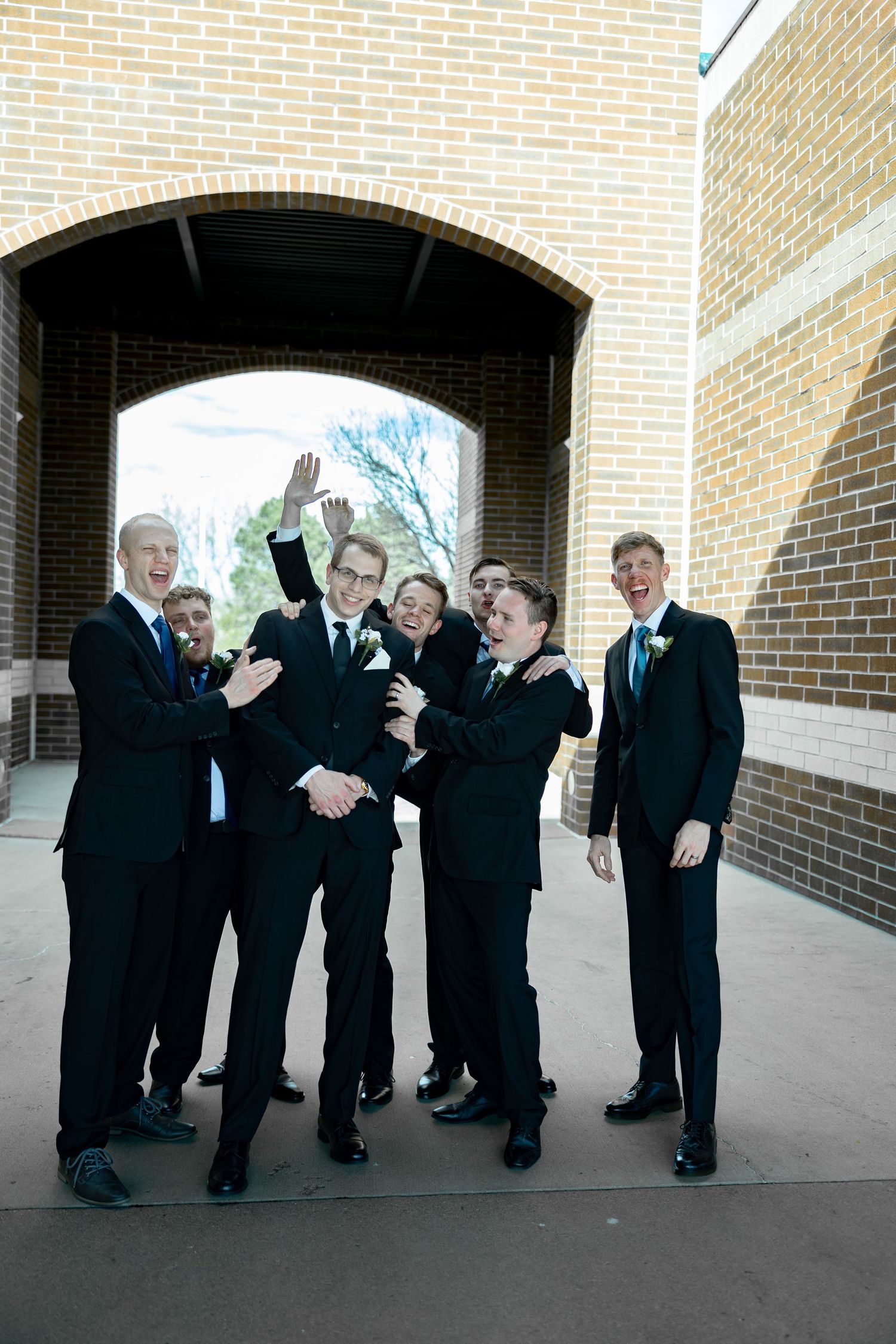 Groomsmen in black suits with teal ties celebrating and laughing under a brick archway at a wedding.