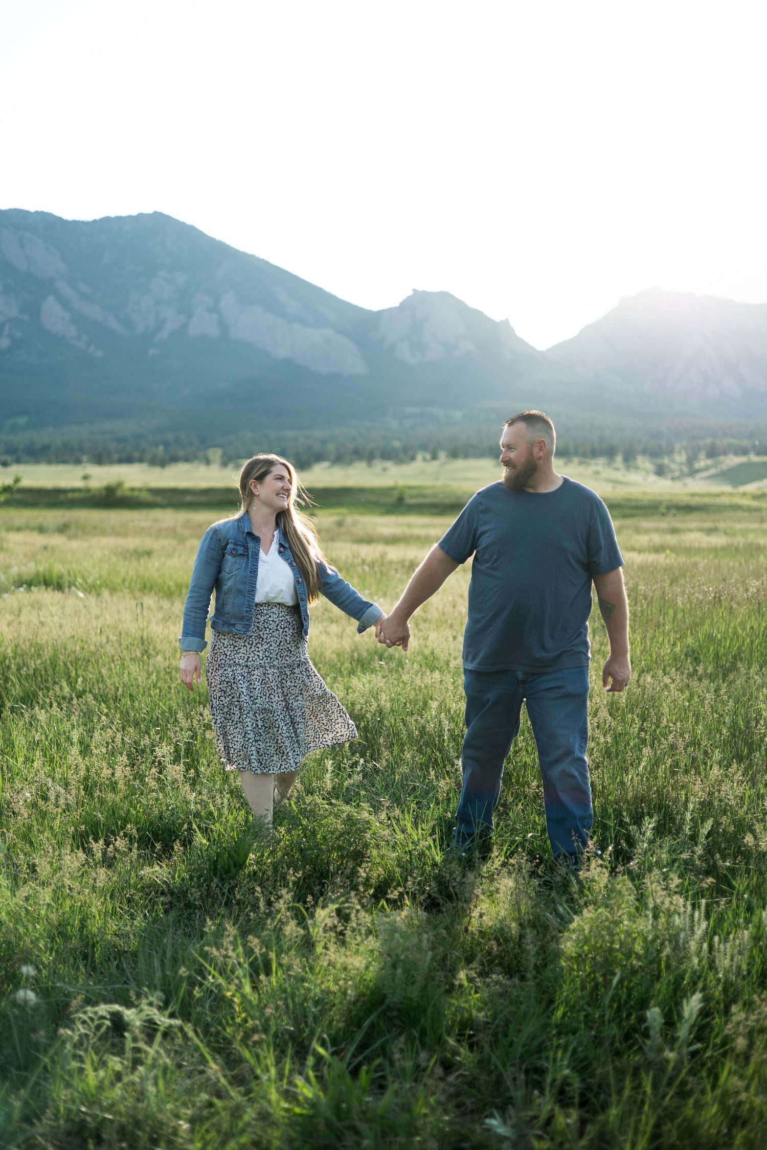 Couple holding hands in a green mountain meadow at sunset, woman in floral skirt and denim jacket, man in blue t-shirt.