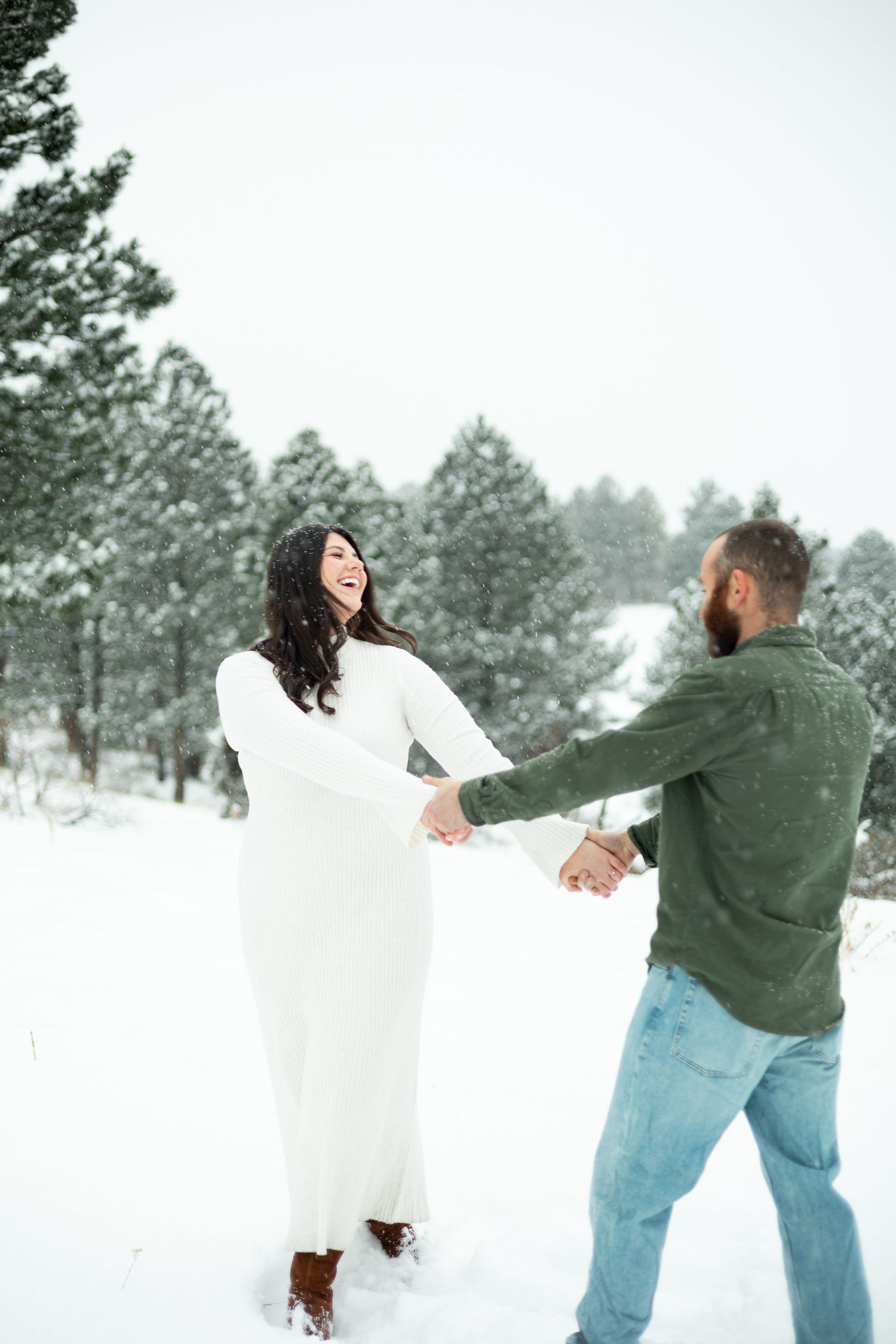 Couple dancing joyfully in snowy winter forest, woman in white dress laughing, man in green shirt holding hands.