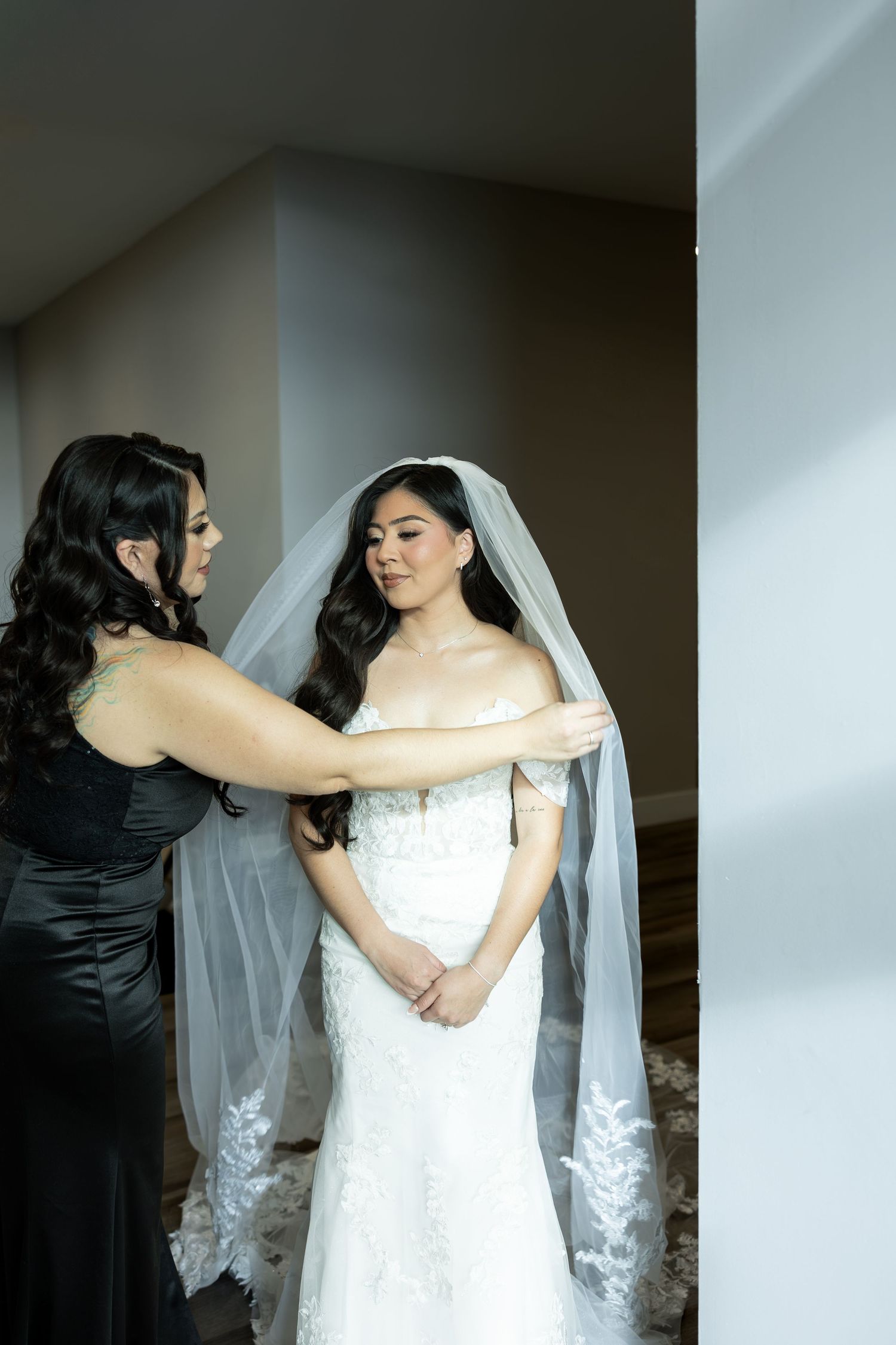 A bride in a white lace dress and veil being helped with final adjustments before the ceremony.