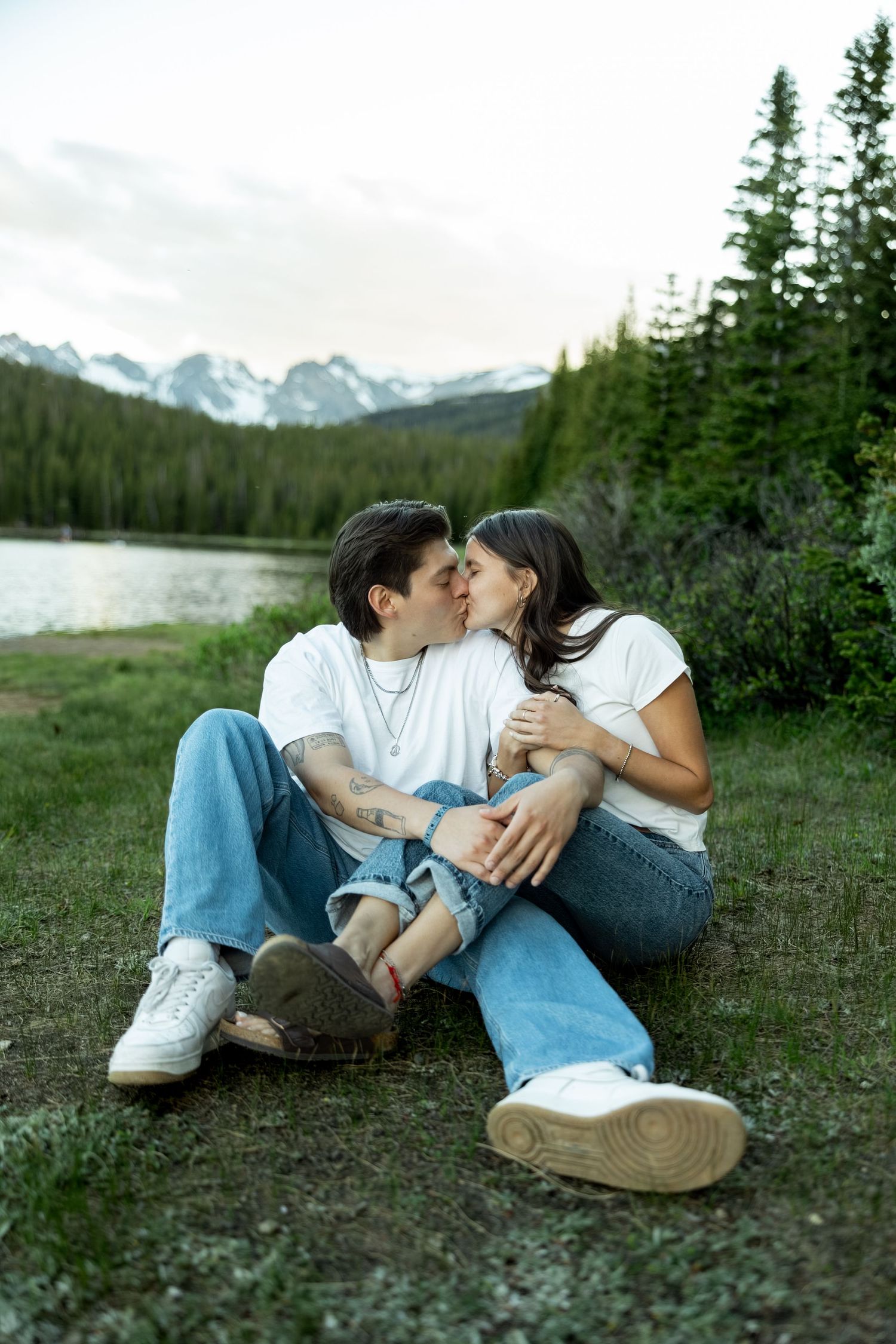 Couple kissing while sitting on grass by a mountain lake surrounded by evergreen trees and snow-capped peaks.