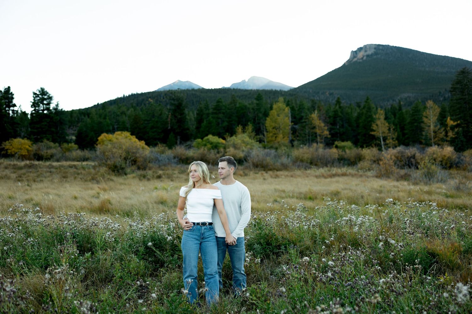 Couple in jeans embracing in a wildflower meadow with evergreen forest and mountain peaks in the background.