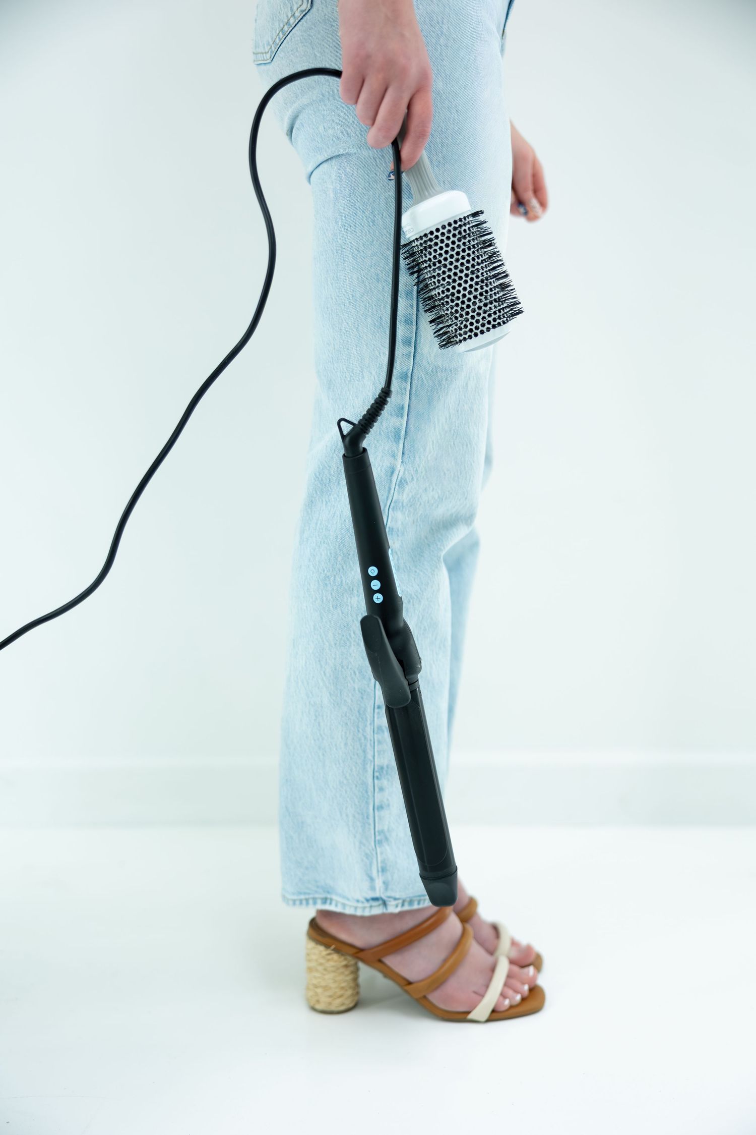 Woman in light blue jeans and cork heeled sandals holding a curling iron and round brush on white background.