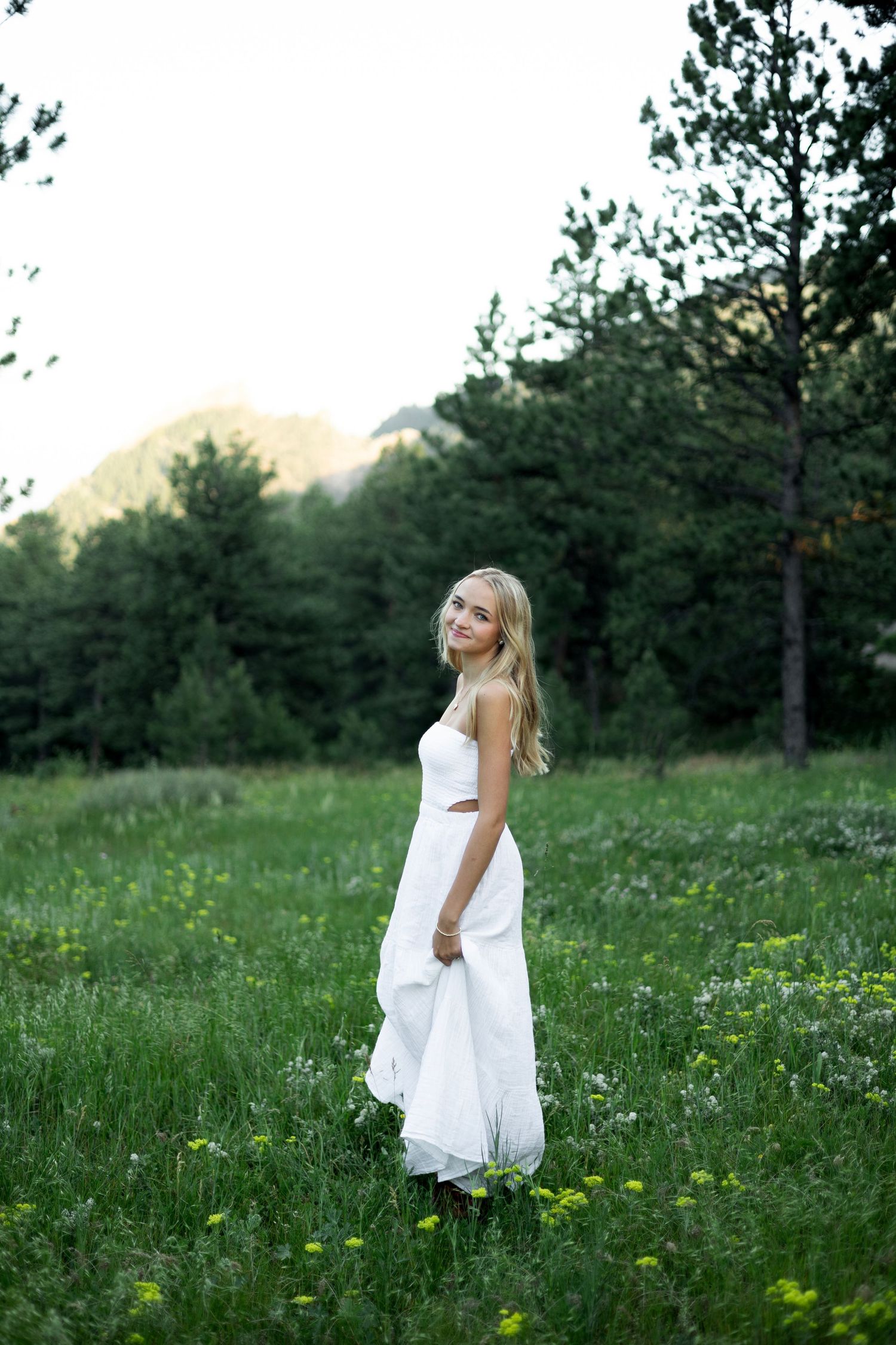 Blonde woman in white dress standing in green meadow with pine trees and mountains in background.