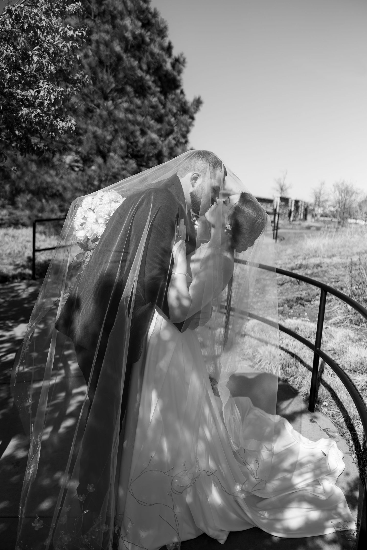 Black and white photo of bride and groom kissing under flowing veil on outdoor staircase.