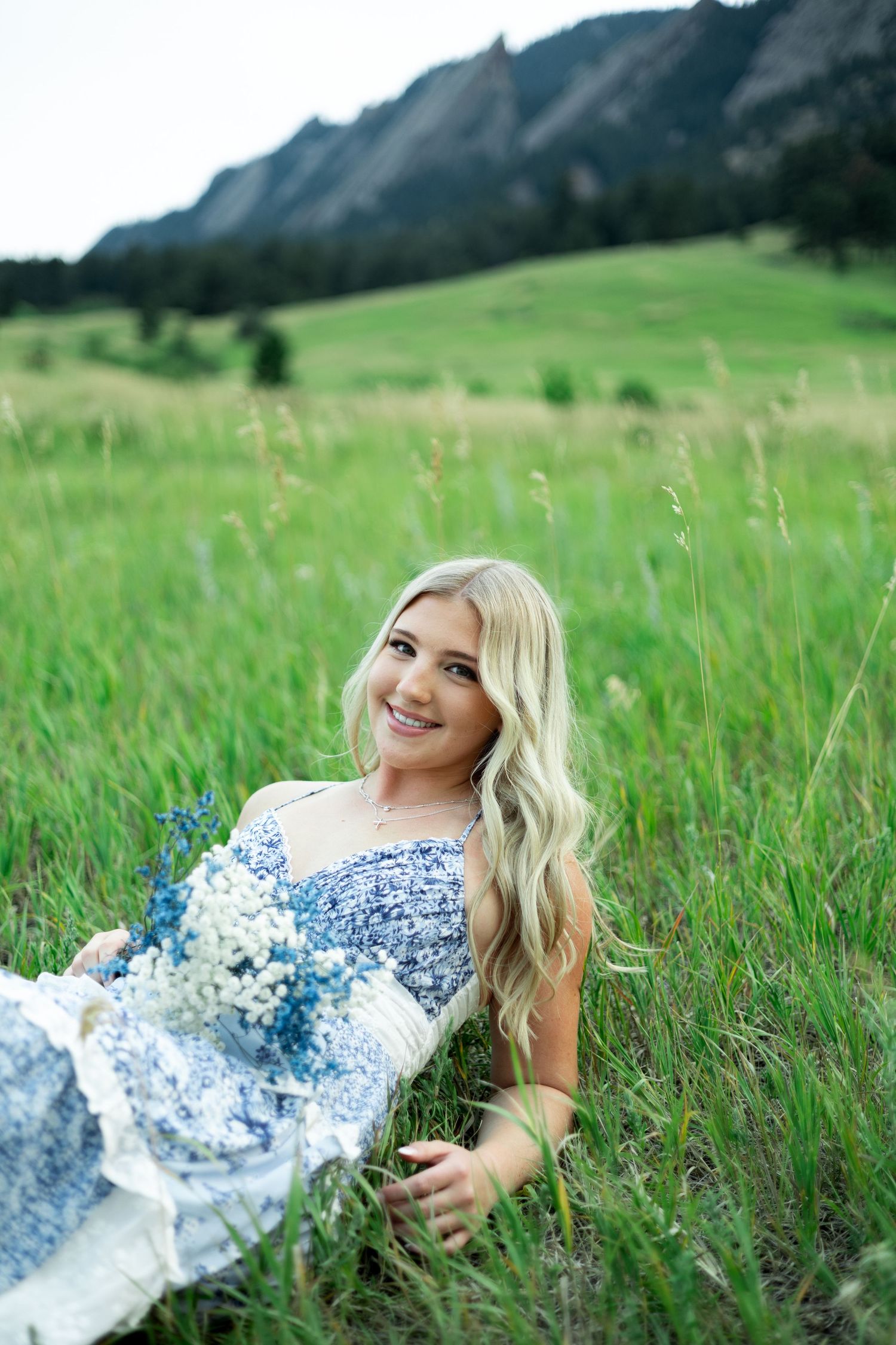 A woman in a blue and white floral dress lays in a green meadow with mountain peaks visible in the background.