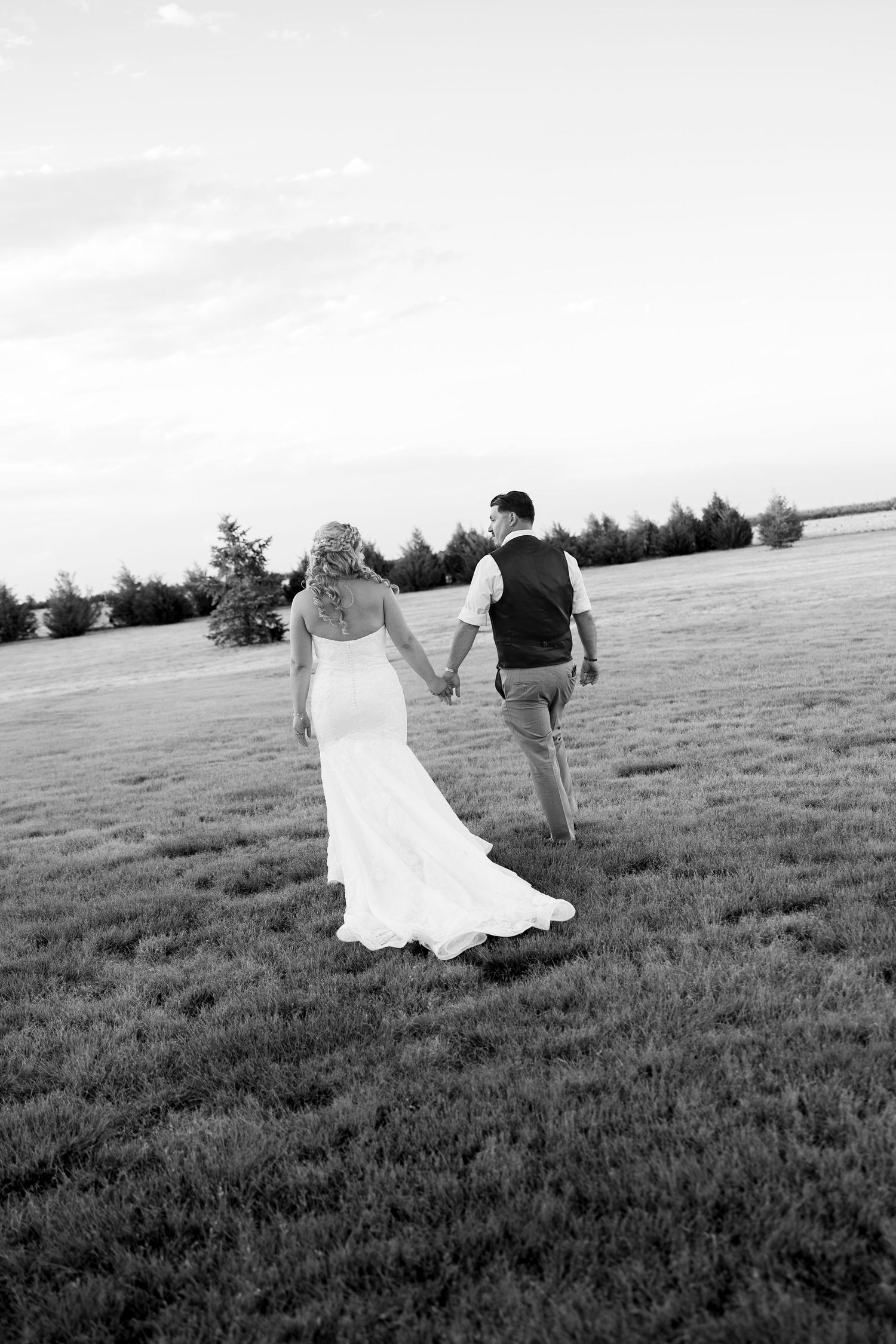 Black and white photo of bride and groom holding hands walking through open field on their wedding day.