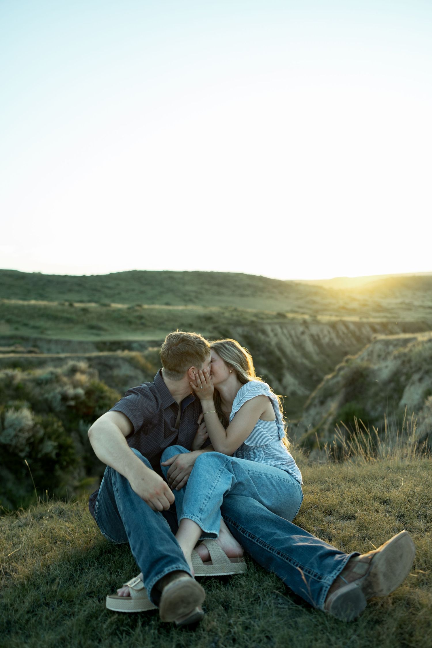 Couple sharing a romantic kiss at sunset in a scenic grassy landscape with rolling hills in the background.